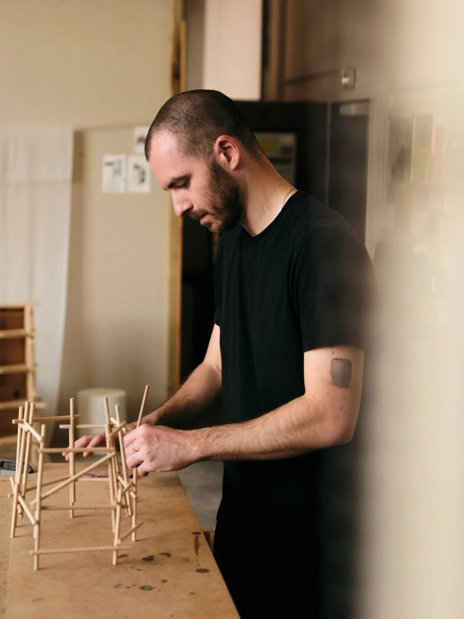 A man with a beard and short hair standing at a table, working on a wooden structure or model.