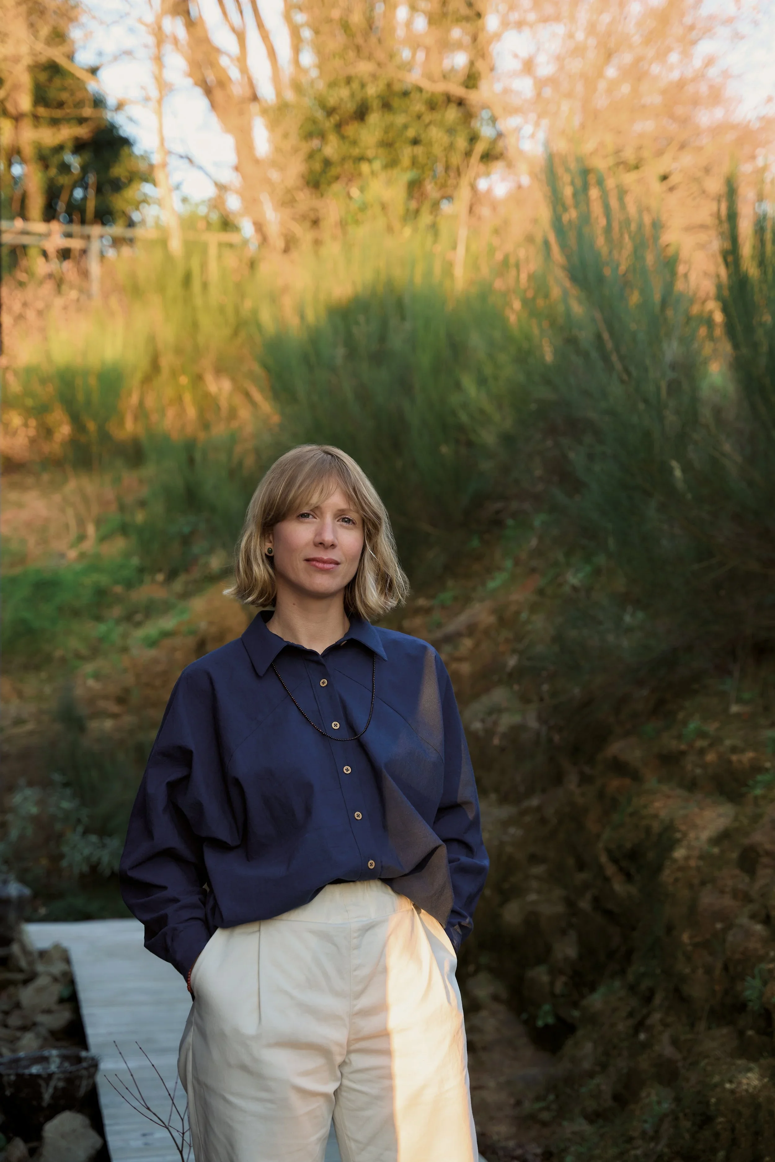 A woman stands outdoors in a natural setting with trees and greenery in the background, wearing a navy blue blouse and beige pants.