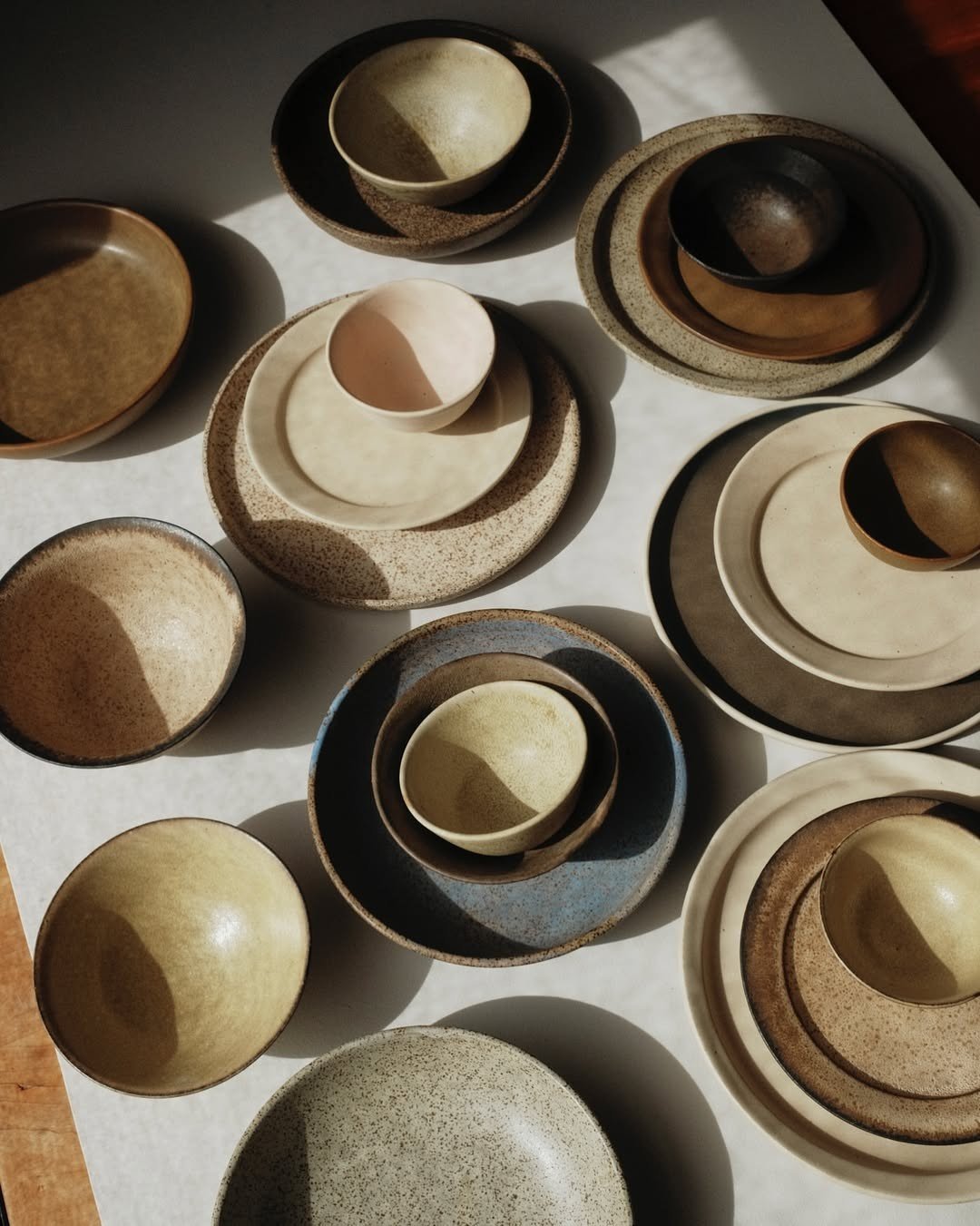 Collection of ceramic bowls and plates in earth tones arranged on a white surface, with shadows cast by sunlight.