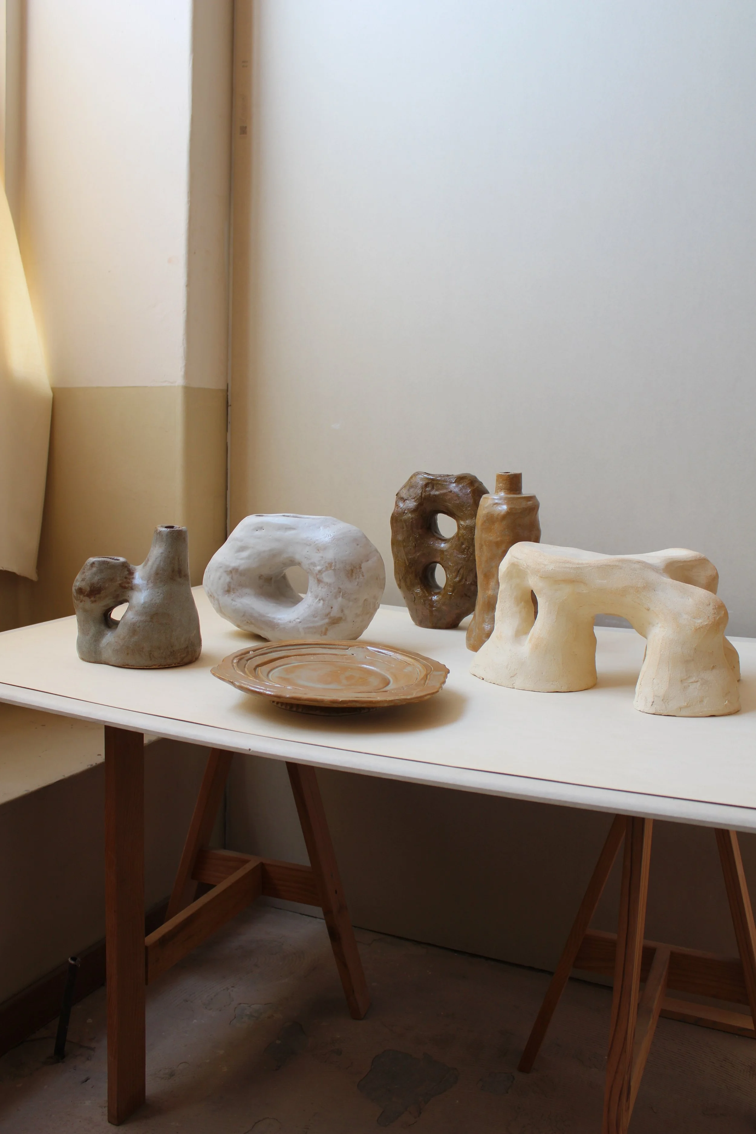 Collection of ceramic art pieces on a white table, with some in the shape of abstract figures and others as plates, in a minimalistic room.