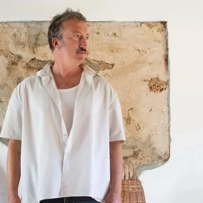 A man with gray hair, mustache, and light skin wearing a white short-sleeve shirt standing in front of a distressed wall with peeling paint, looking to the side.