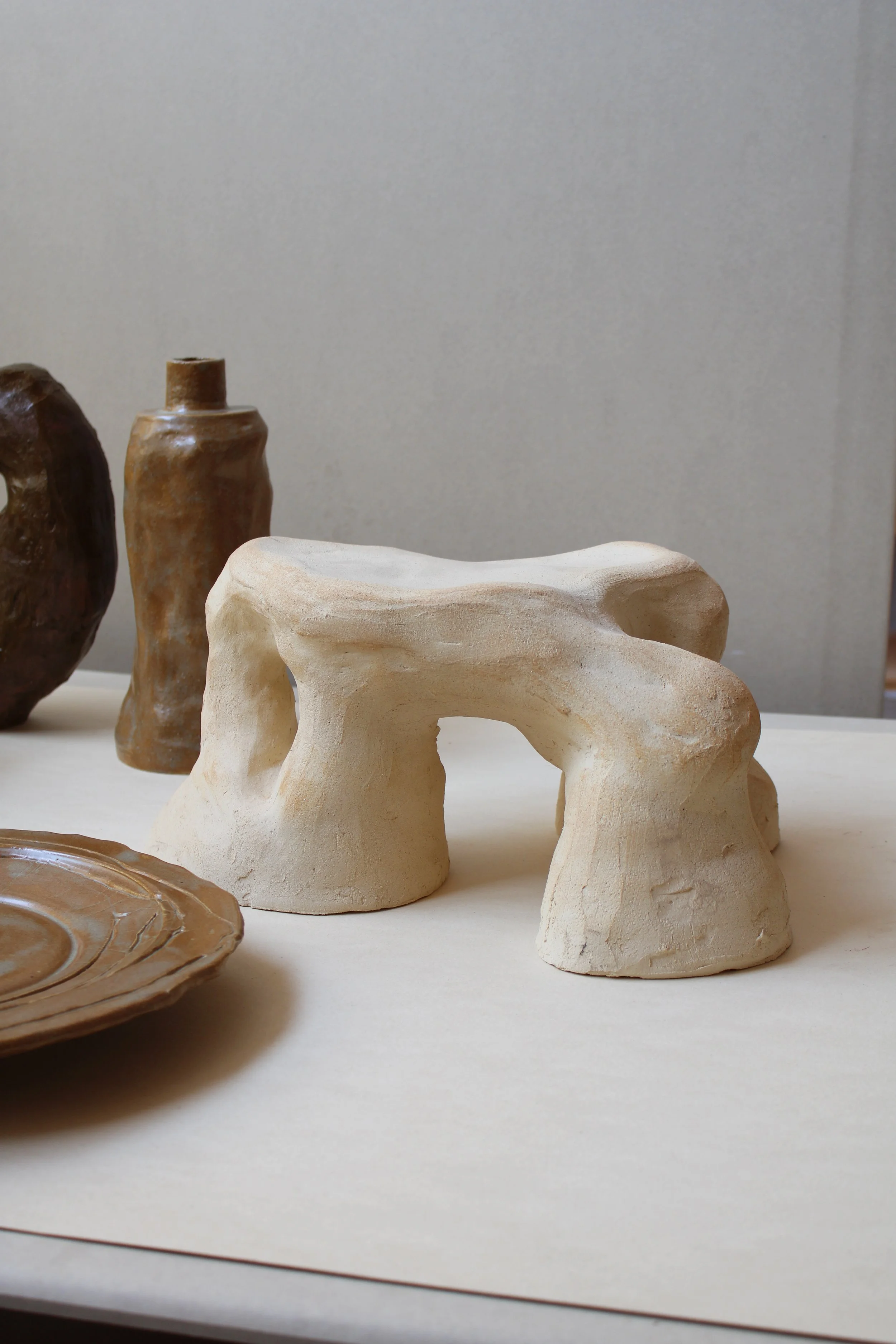 Arrangement of various pottery pieces, including a single brown bottle, a multi-hole pot, and a white ceramic piece with arch-shaped structures, on a white surface with a pale background.