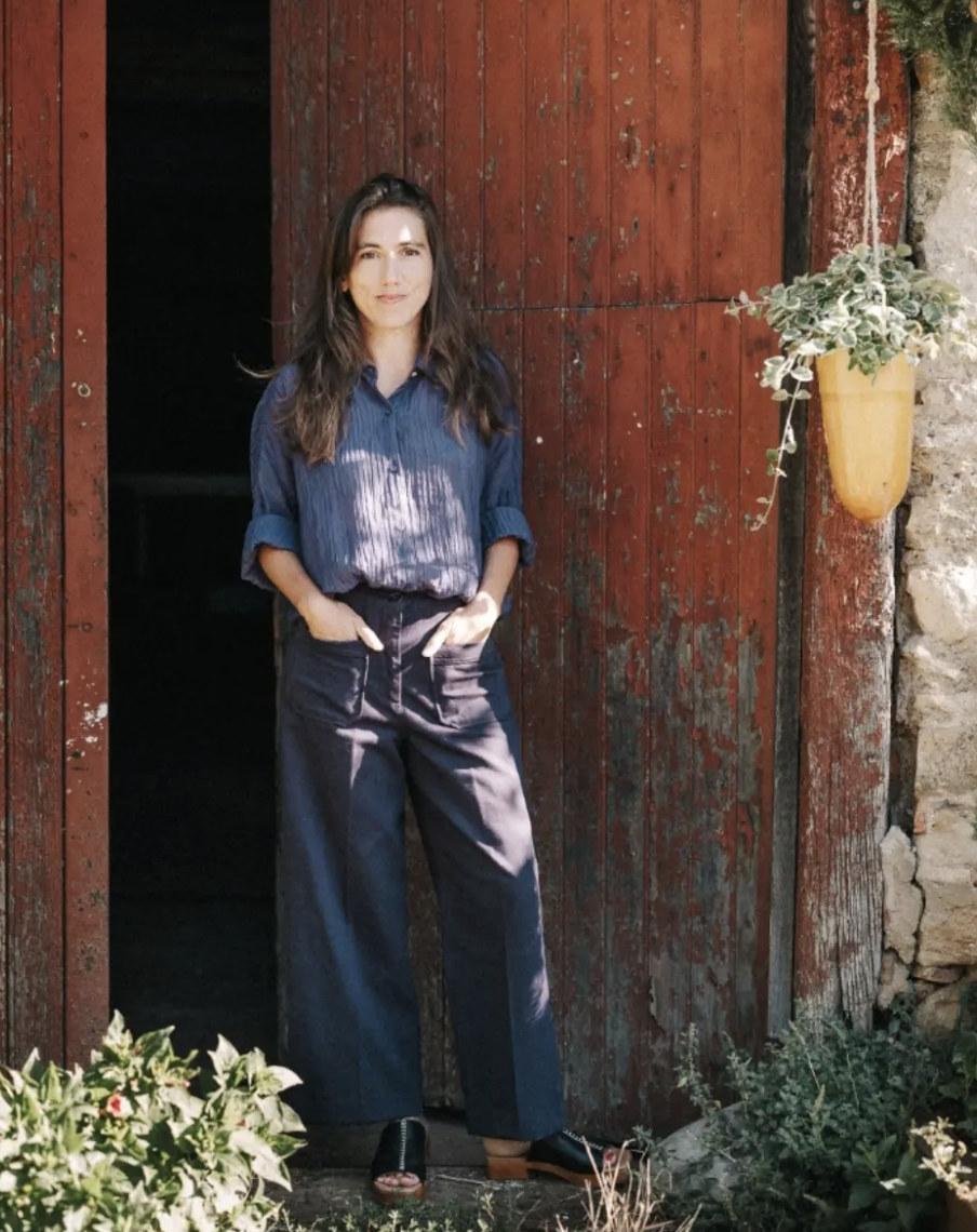 A woman with long brown hair wearing a blue shirt and black pants standing in front of a rustic red wooden door with a hanging flower pot on the side.