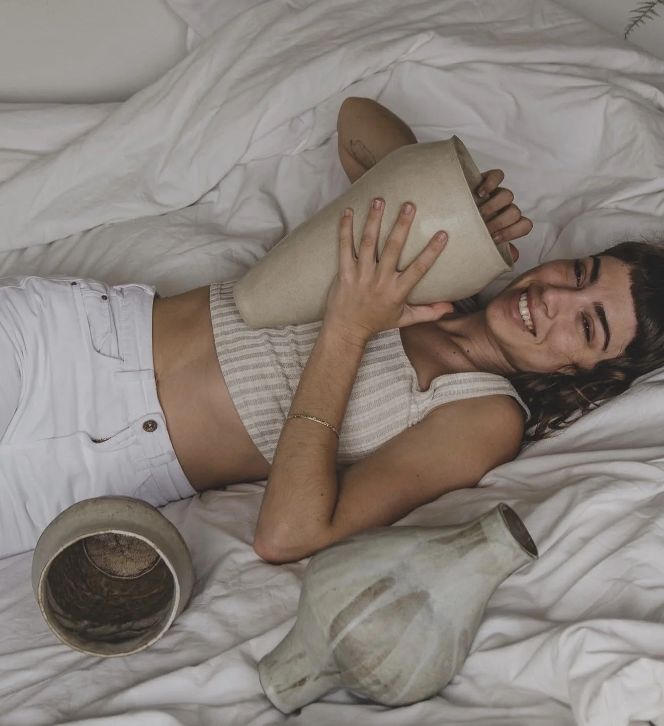 Woman lying on bed holding a large vase, smiling, surrounded by empty ceramic vases.