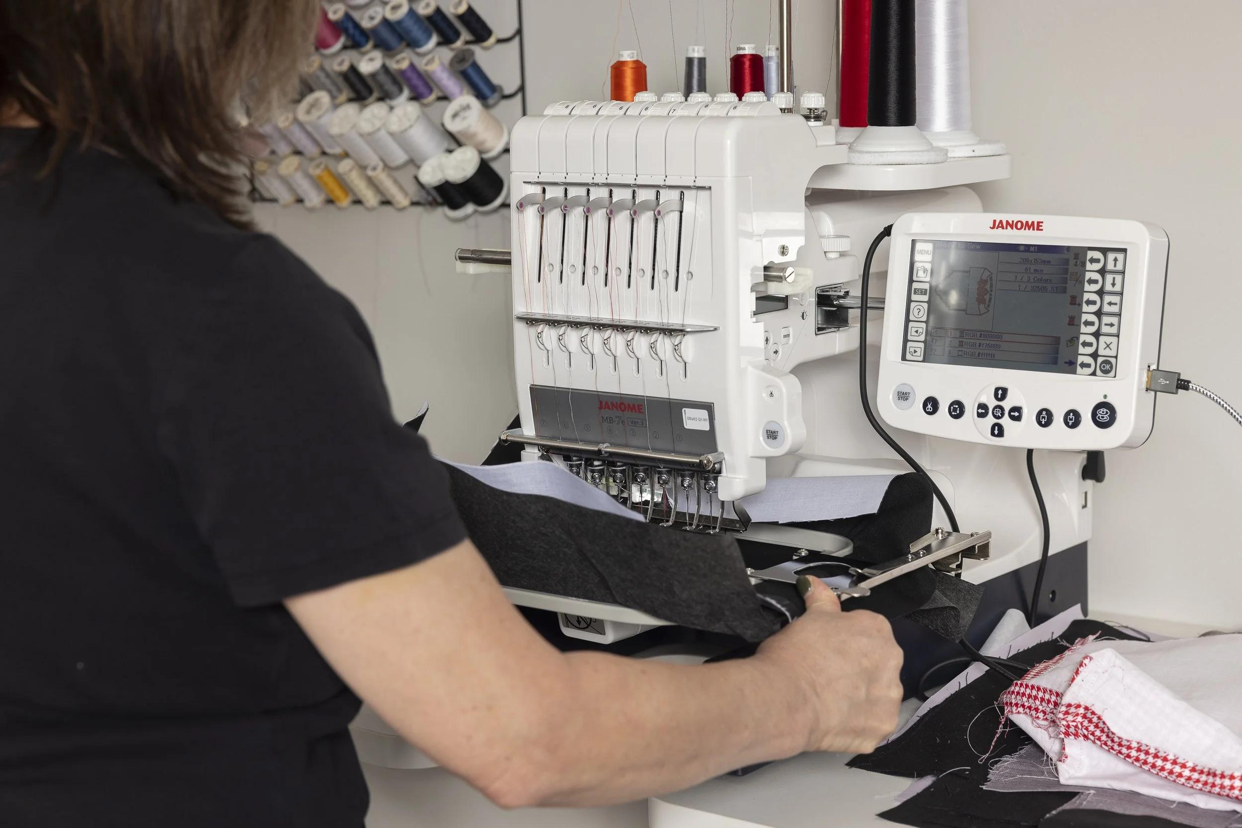 A close-up of a person operating a computerized embroidery machine sewing fabric with black, white, and red thread, in a sewing workshop.