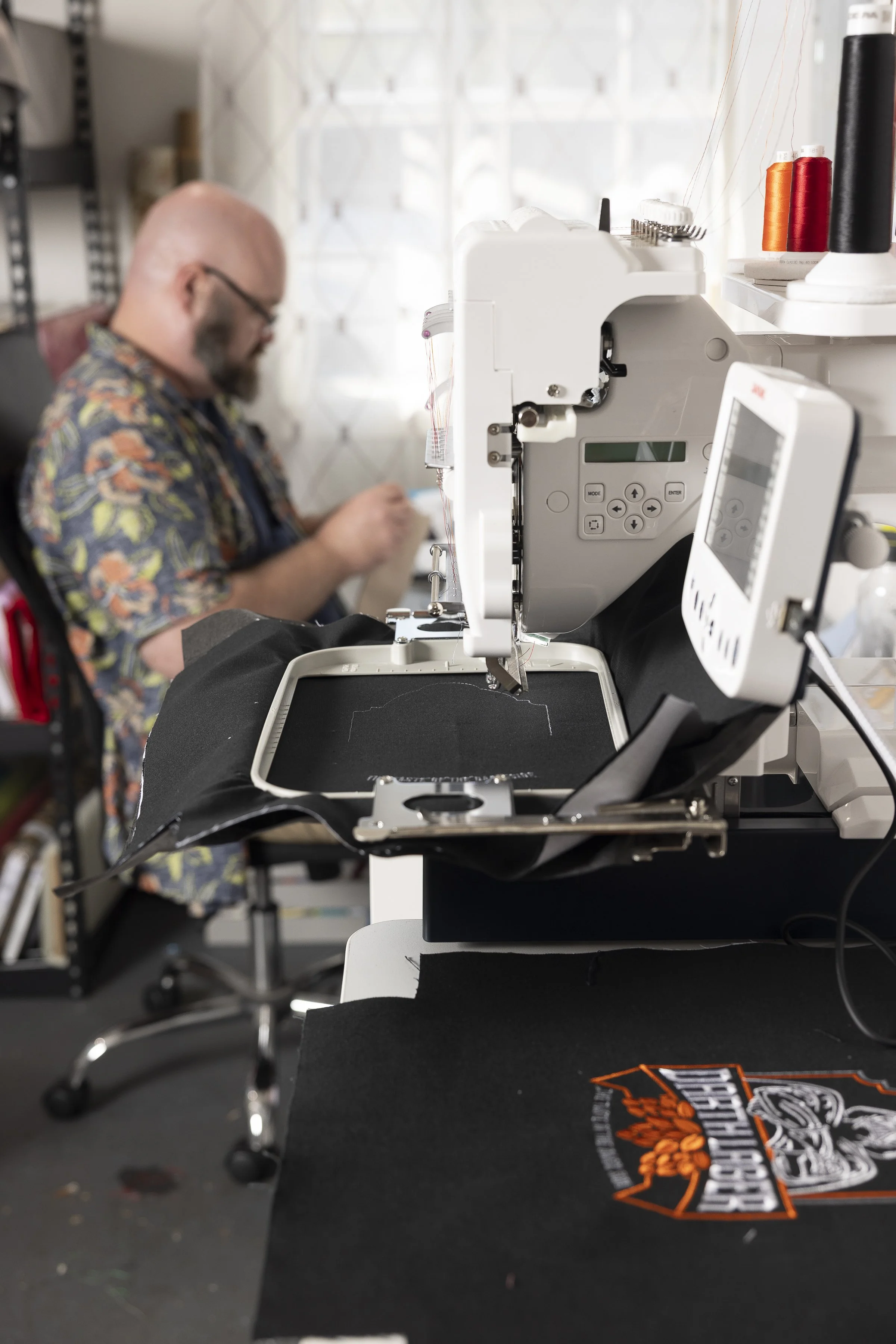 A person sewing on a sewing machine in a workshop, with fabric featuring a Harley-Davidson logo in the foreground.