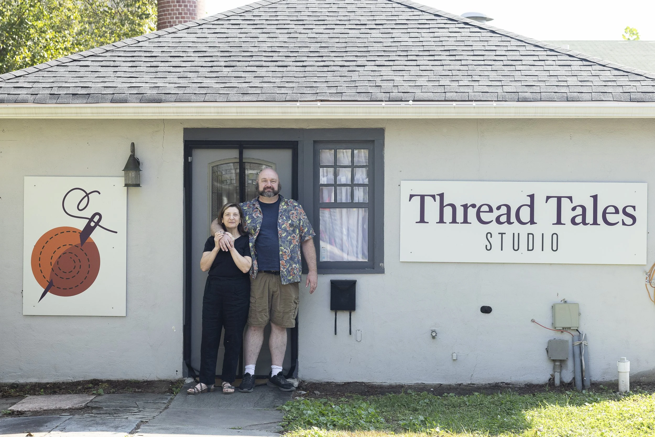 A man and woman standing in front of a building called Thread Tales Studio, with the woman holding the man's arm. The building has a sign on the right with the words 'Thread Tales Studio' and a colorful logo on the left featuring a needle and thread.