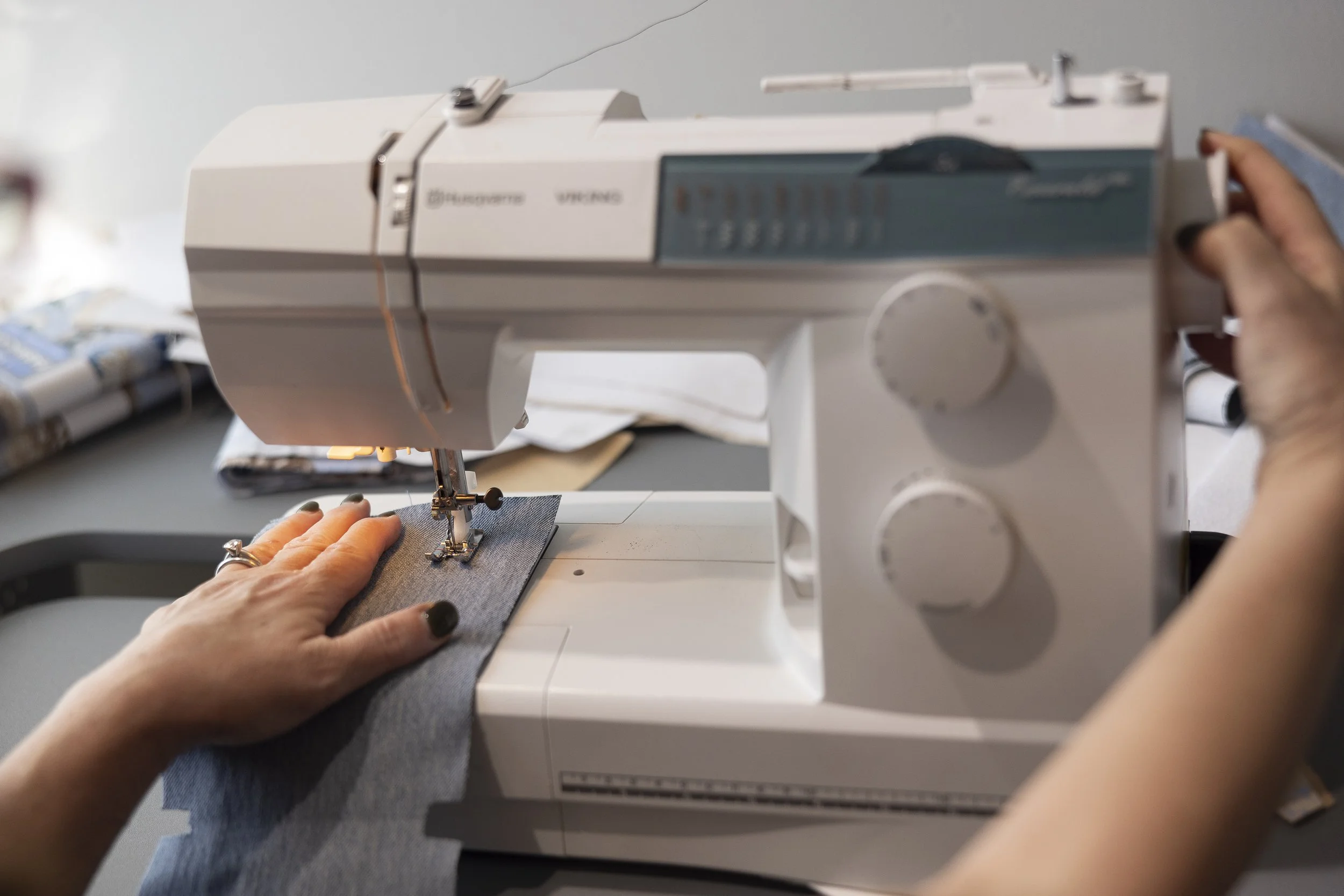 Hands operating a white sewing machine sewing gray fabric.