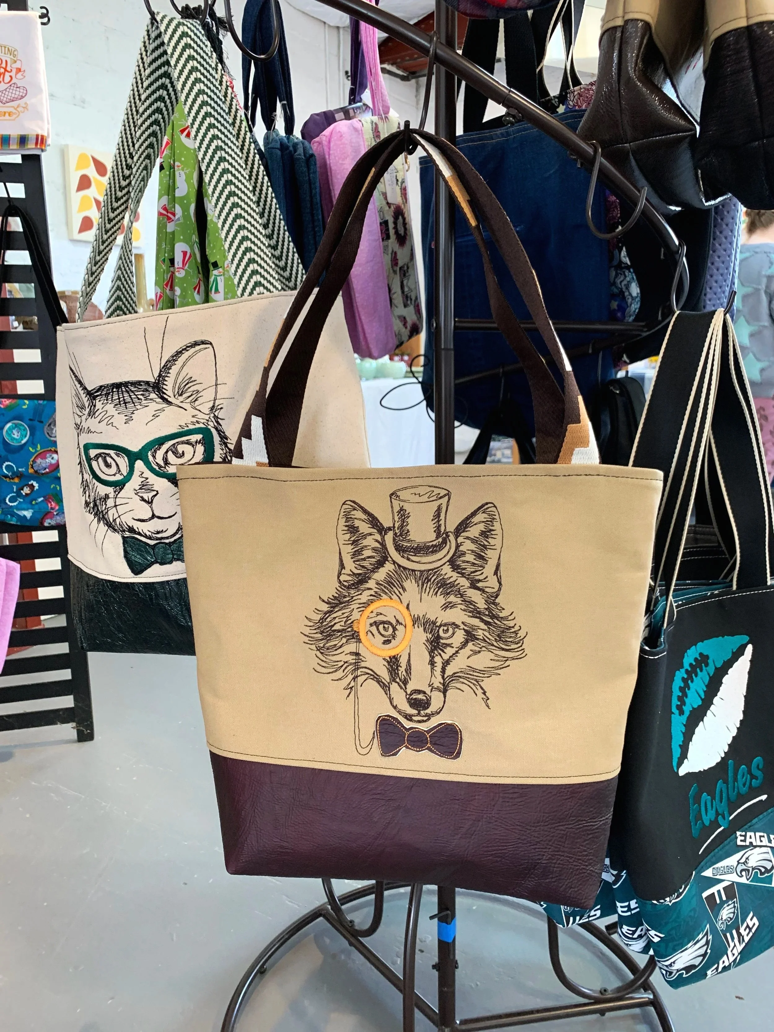 Tote bags with animal illustrations hanging on display. One features a cat with glasses and a bow tie, and the other has a wolf with a top hat and bow tie.