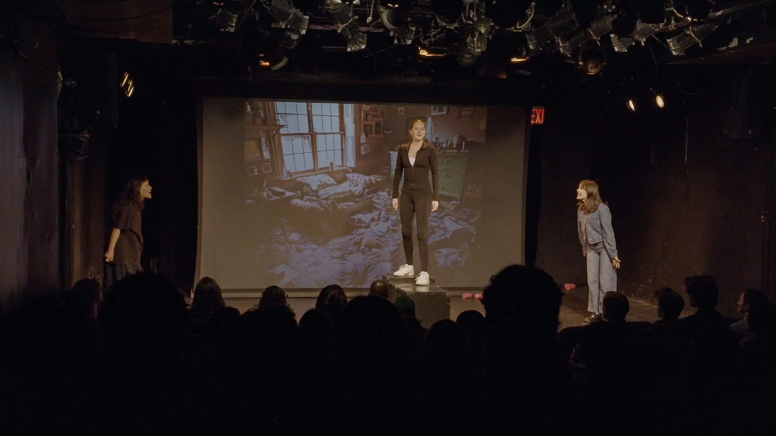 Three women perform on a stage with a large screen in the background displaying an image of a messy room.