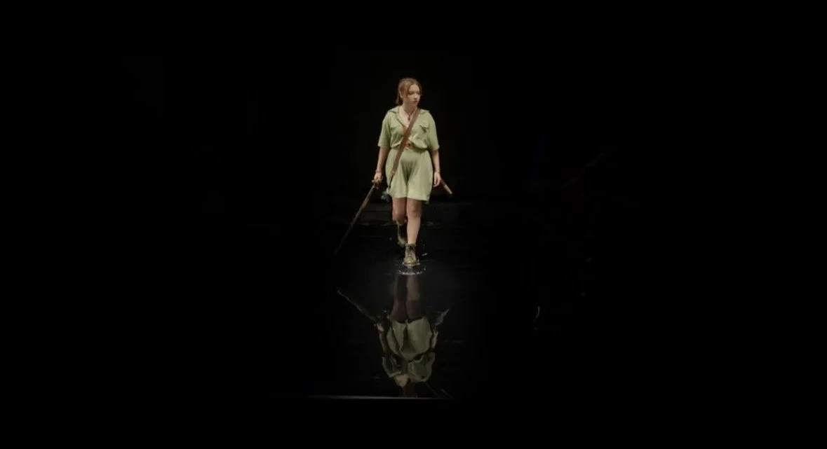 Young woman in green dress walking on a dark reflective surface, holding a stick, with her reflection visible on the surface.