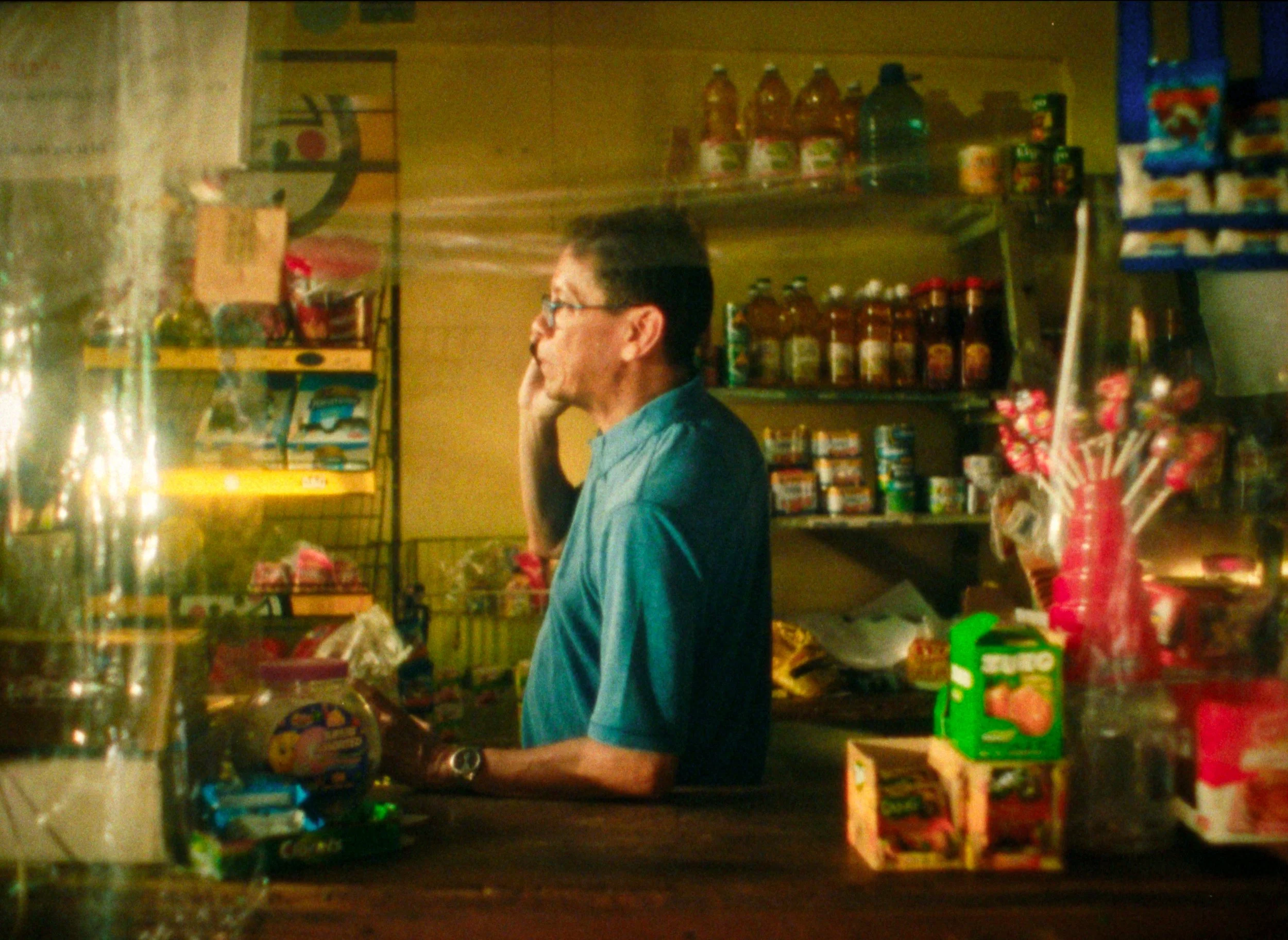 A man in glasses and a blue shirt standing behind a store counter inside a small shop, with various food and beverage products displayed on shelves around him.