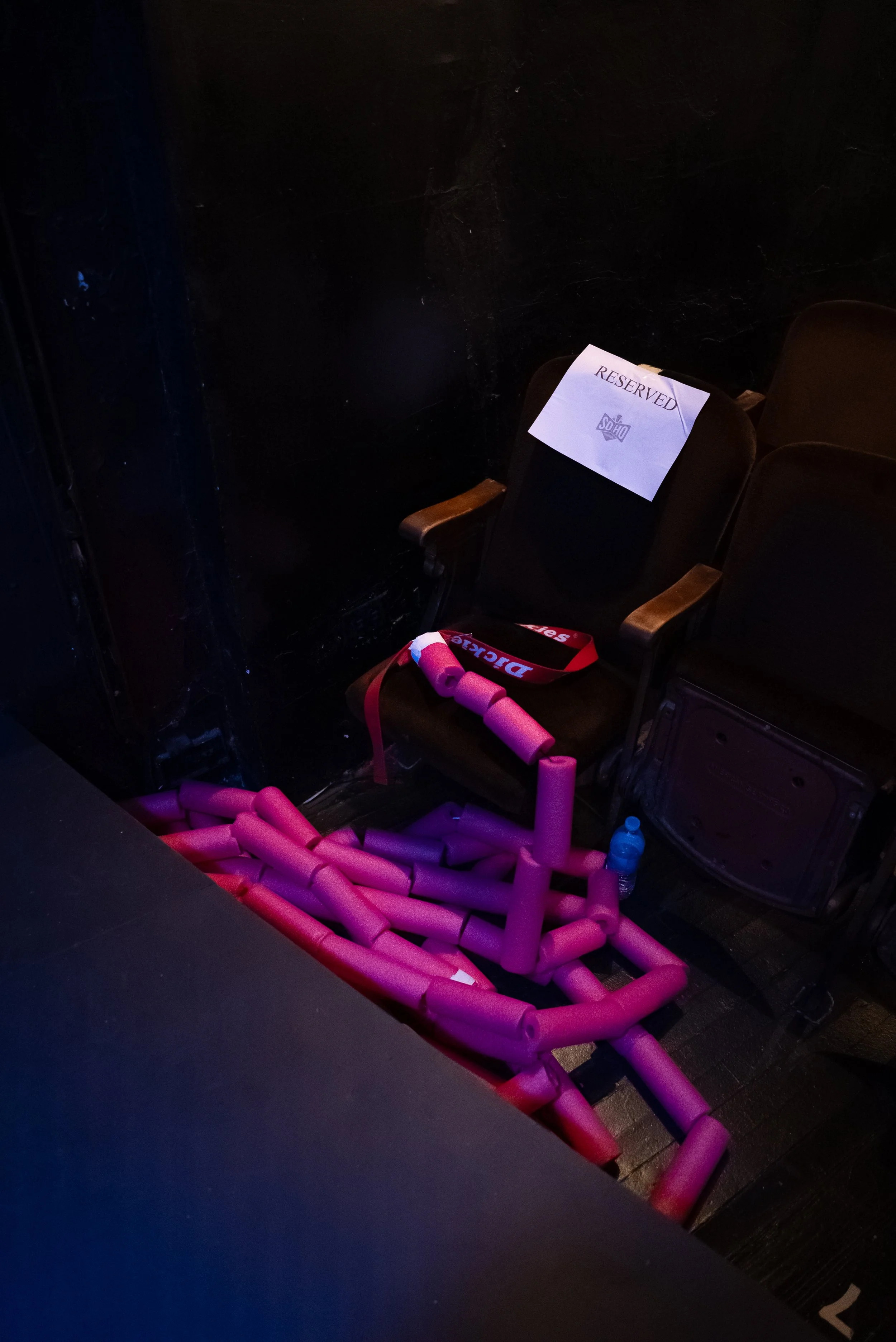 A reserved seat in a dark theater with a sign on it, pink foam pool noodles on the floor, and a water bottle next to the seat.