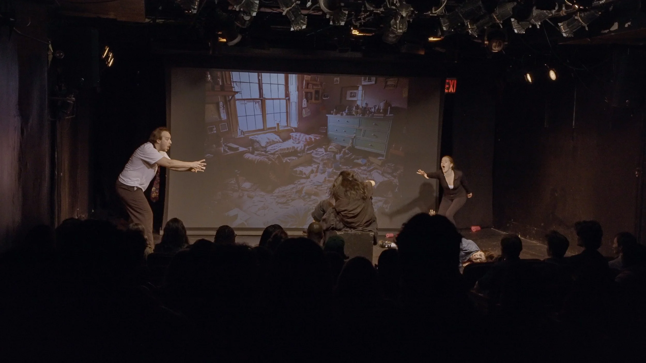 Theater troupe performing on stage with a large projected image of a messy bedroom in the background. Three actors are visible, actively engaged in a scene.