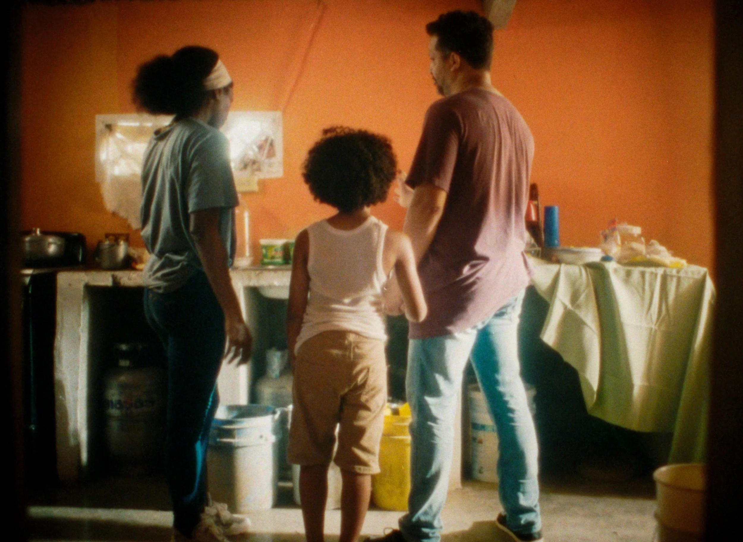 Four people stand in a kitchen with an orange wall, two children and two adults, seen from behind, engaged in conversation or activity.
