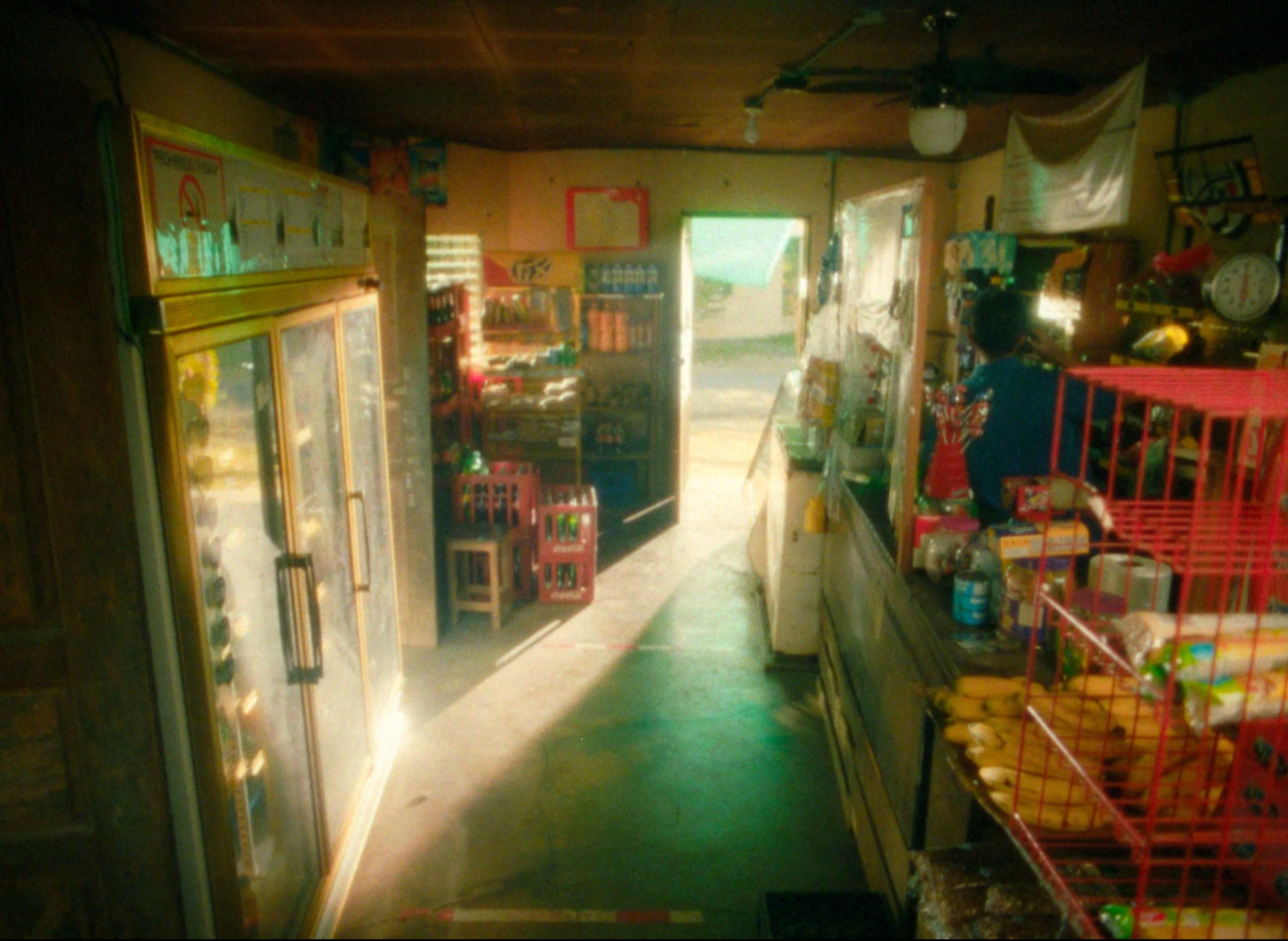 Interior of a small convenience store with shelves stocked with snacks and drinks, a customer at the counter, and an open door leading outside.