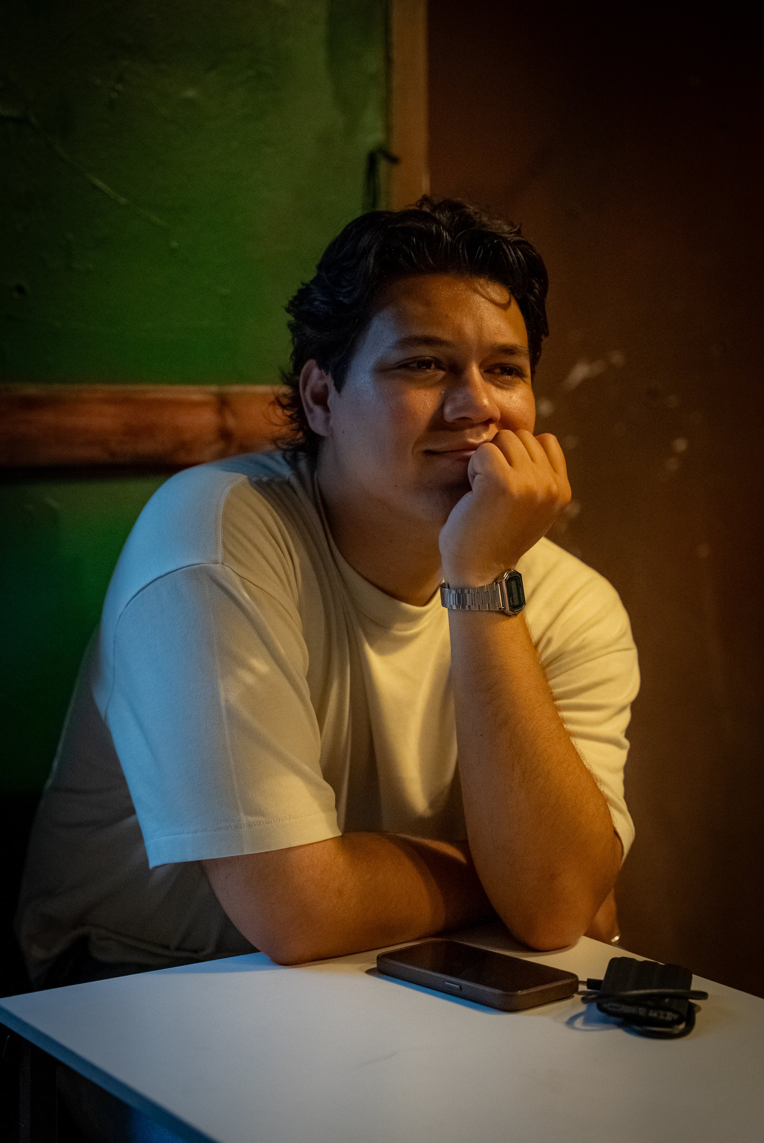 A young man with dark hair, wearing a beige t-shirt and silver watch, resting his chin on his hand, sitting at a table with a smartphone and charger, in a dimly lit room with green and brown walls.