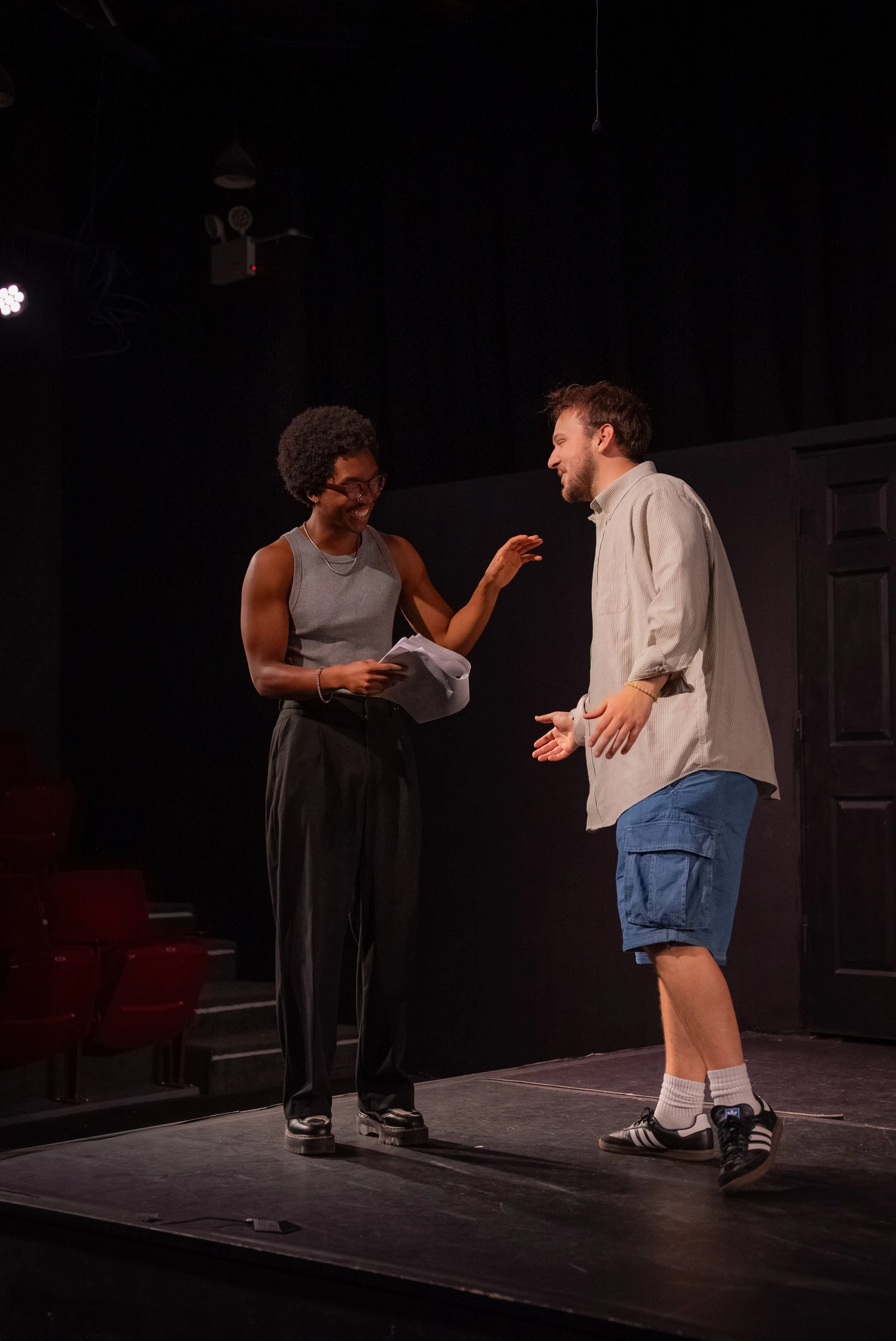 Two actors rehearsing on stage with black background, red seats visible on the side, one with curly hair and gray tank top, the other in a light shirt and shorts, engaged in a conversation or scene.
