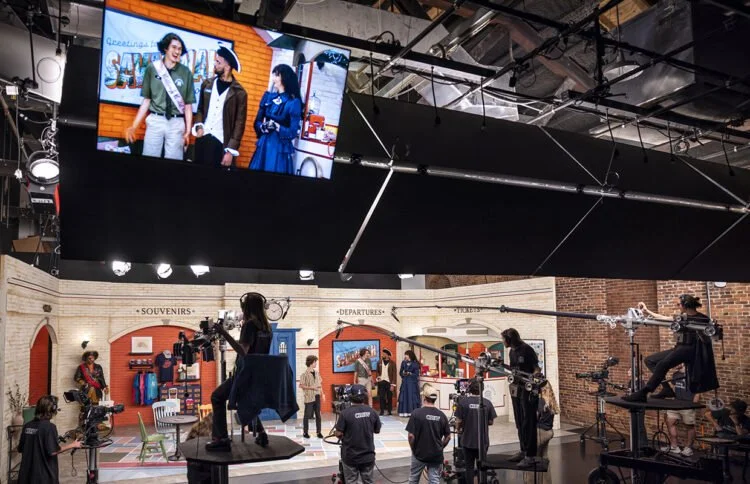Behind the scenes of a TV or film production set with a large monitor displaying three people, set pieces labeled 'Souvenirs' and 'Departures,' and crew members operating cameras and equipment in a studio.