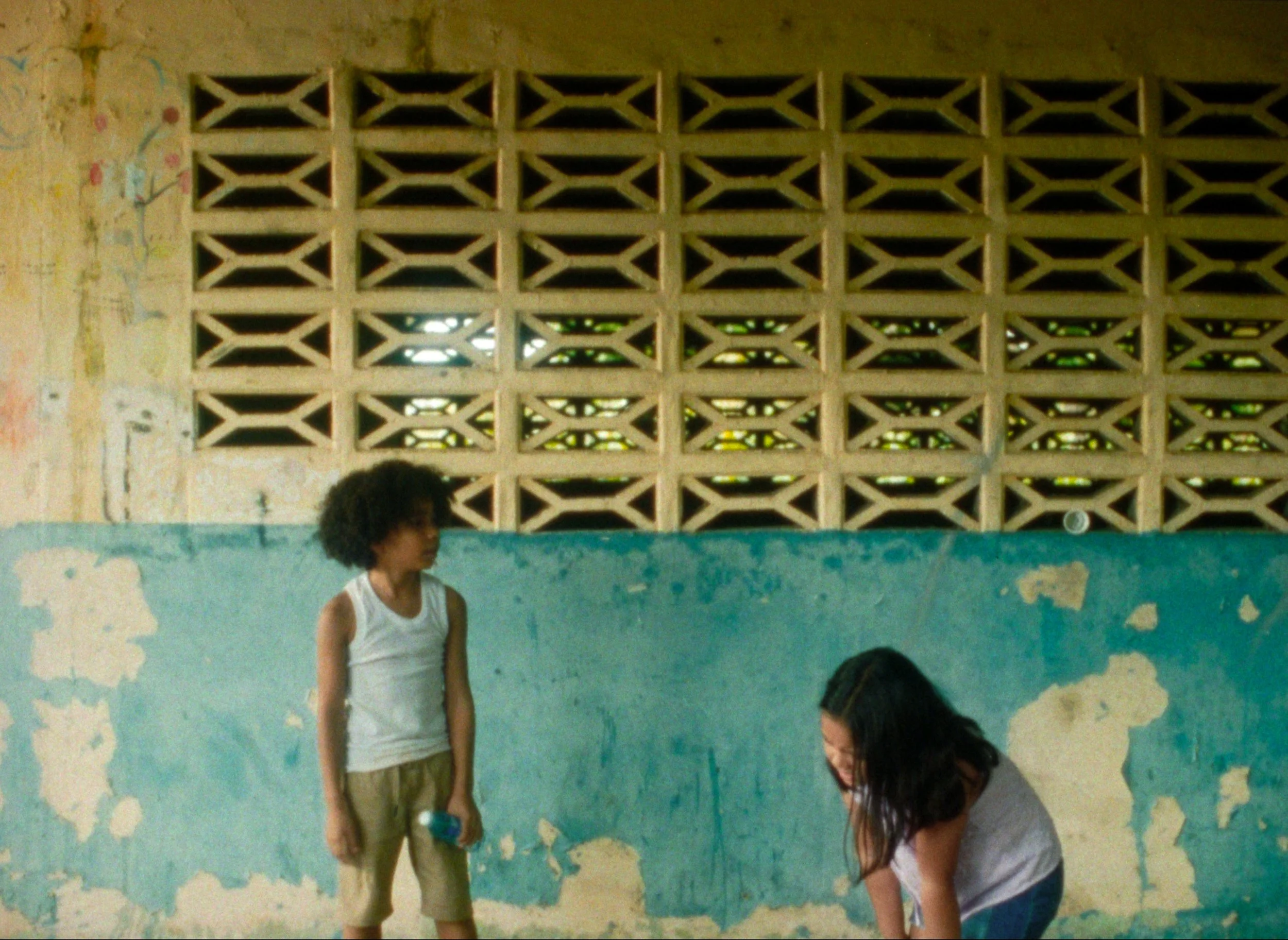 Two children, one girl with curly hair standing and a girl with long straight hair squatting, in front of a weathered wall with peeling paint and a decorative brick wall.