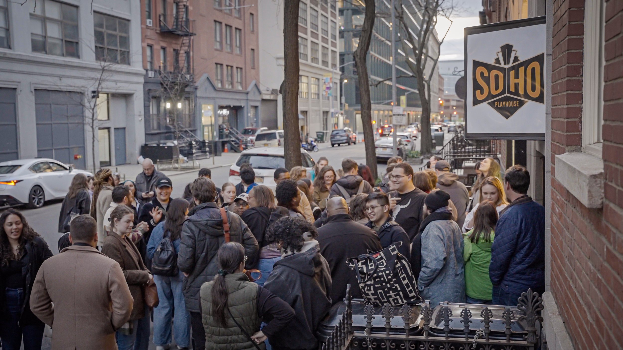 Crowd of people gathered outside SOHO Playhouse on a city street, with cars parked and driving by in the background.