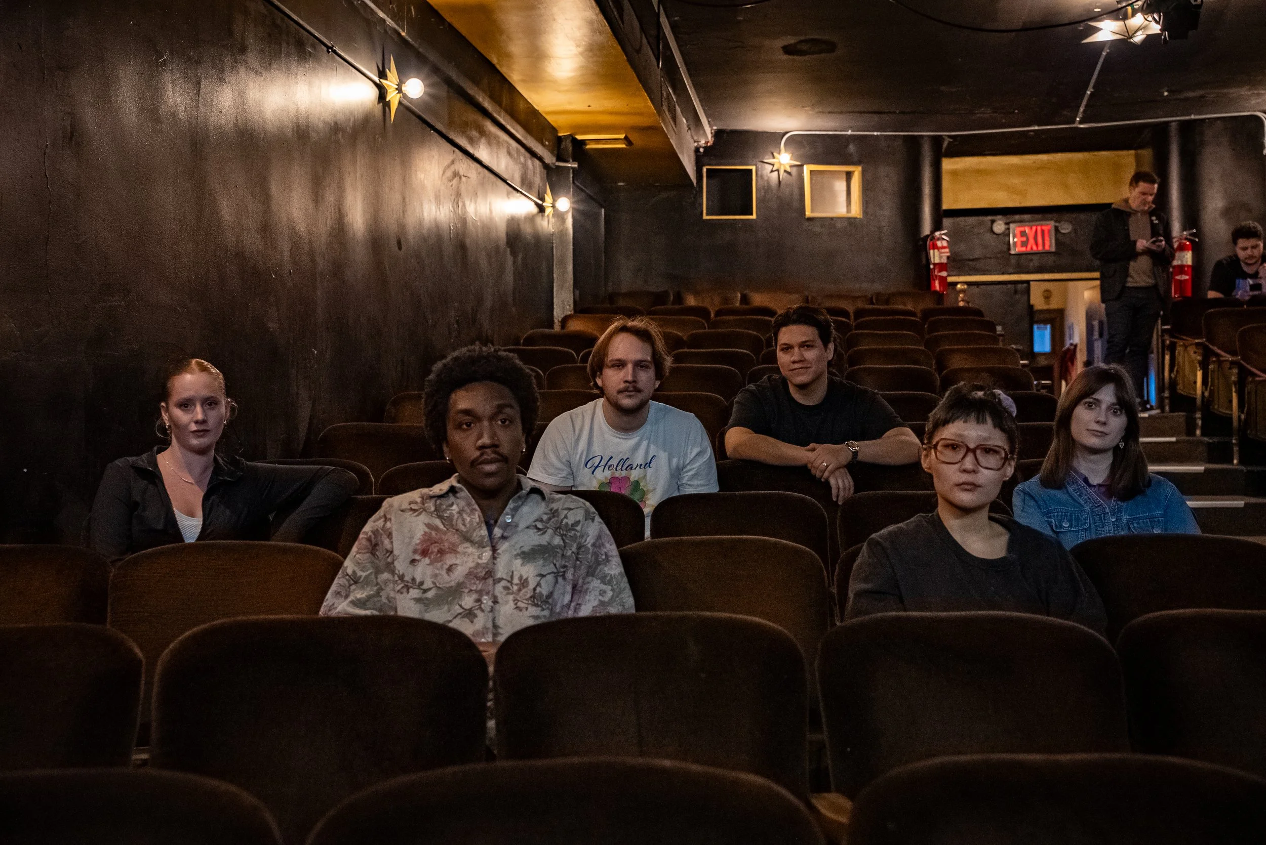 Six people sit in an empty movie theater, six chairs in front of them, dark walls, some decorative lights, and an exit sign in the background.