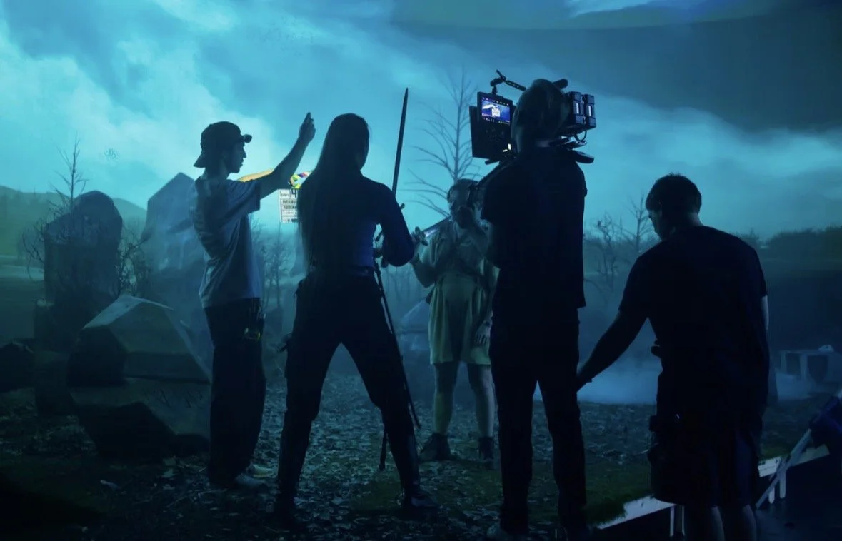 Behind the scenes of a film or TV scene being shot outdoors with crew members, equipment, and a person in costume, against a blue-toned cloudy sky and eerie landscape.