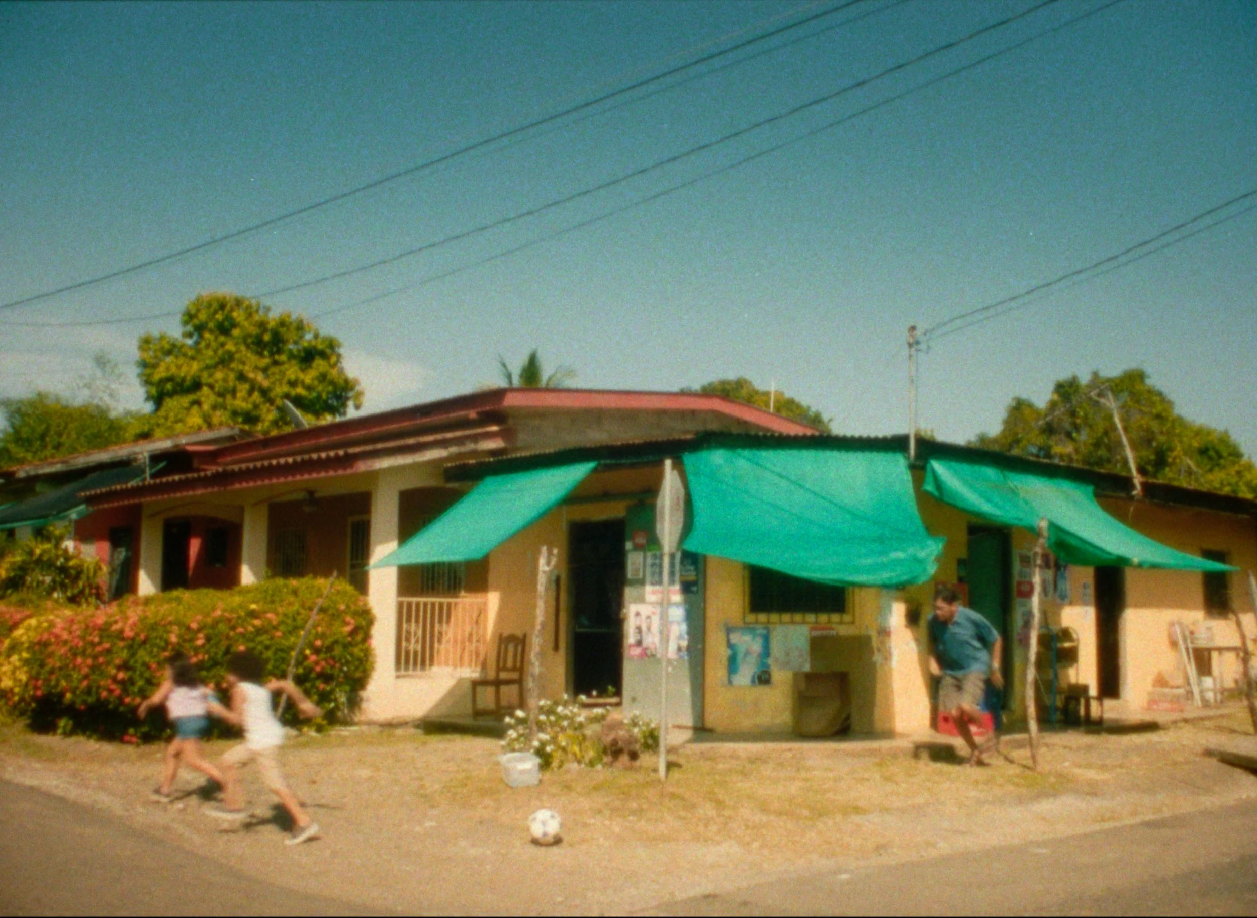 A house with a red roof and yellow walls, with green shade cloths hanging over the front and side, and two children playing on the street in front.