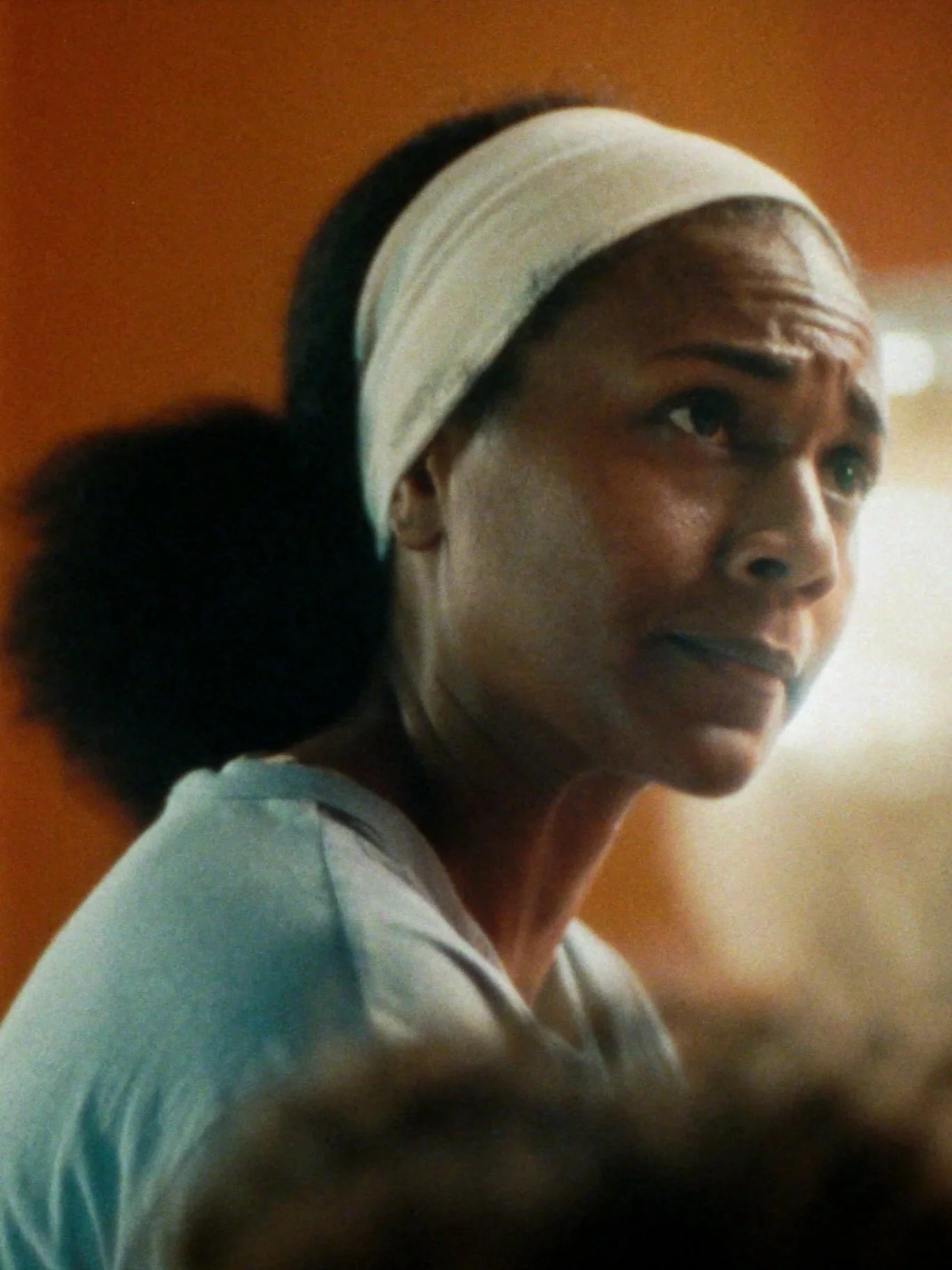 A woman with a light-colored headband, dark hair in a ponytail, and wearing a light-colored shirt, looks to her right with a thoughtful expression.