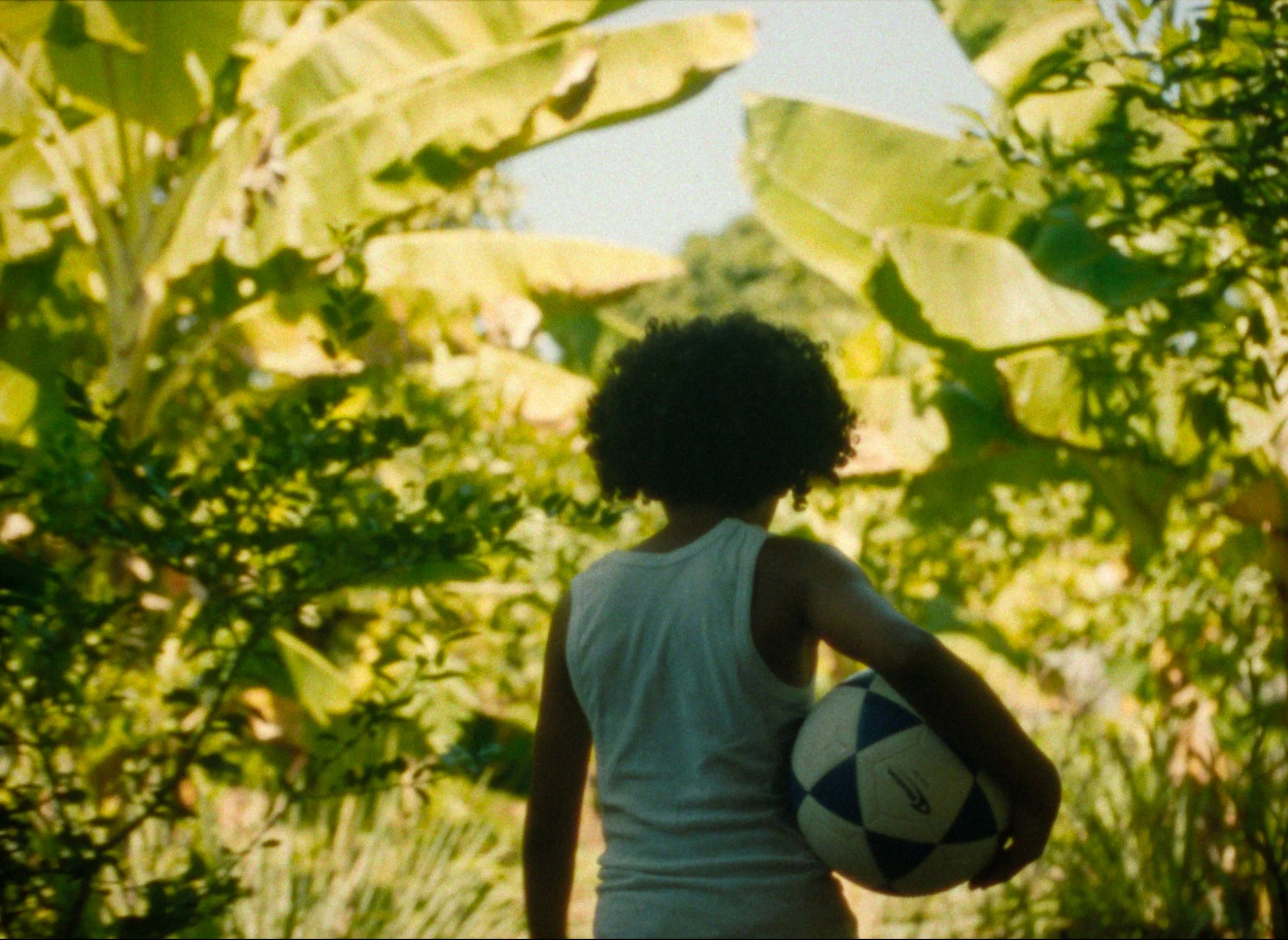 A person with curly hair, wearing a white sleeveless shirt, standing outdoors among greenery, holding a soccer ball.