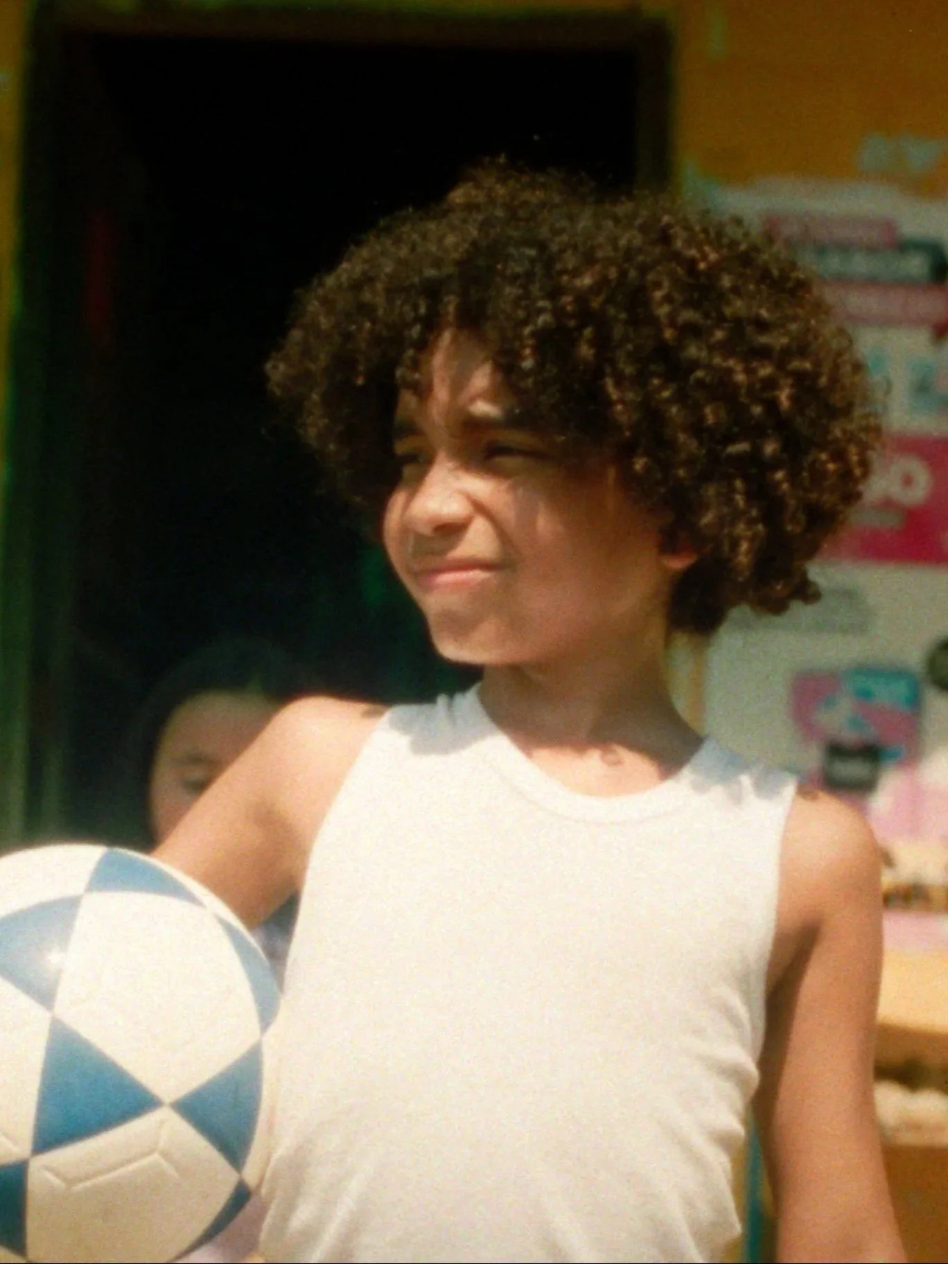 A young boy with curly hair in a white sleeveless shirt holding a soccer ball. El Alma de la Sed