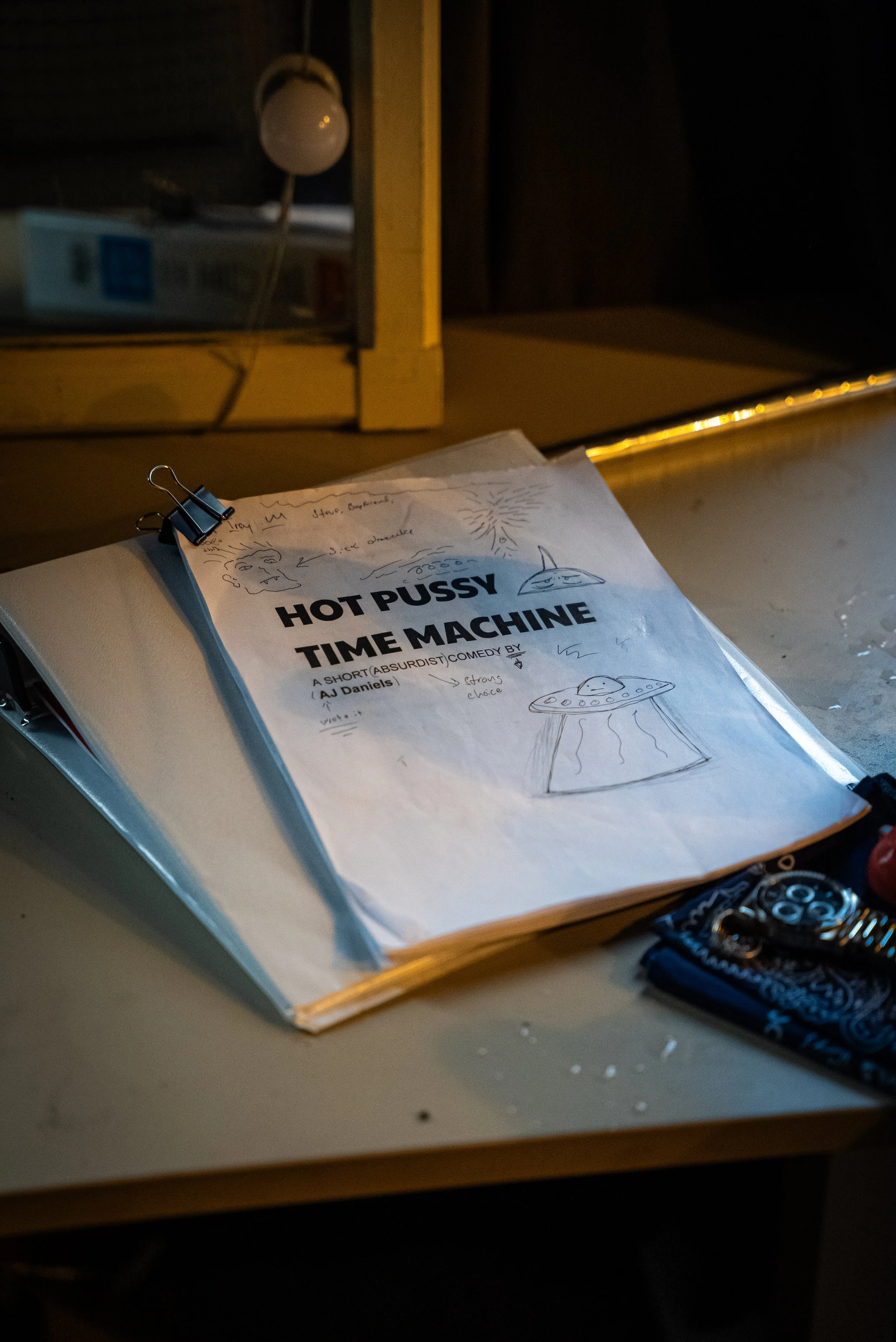 A script titled 'Hot Pussy Time Machine' with handwritten notes and sketches of UFOs and aliens, clipped to a binder on a desk with a watch and bandana nearby.