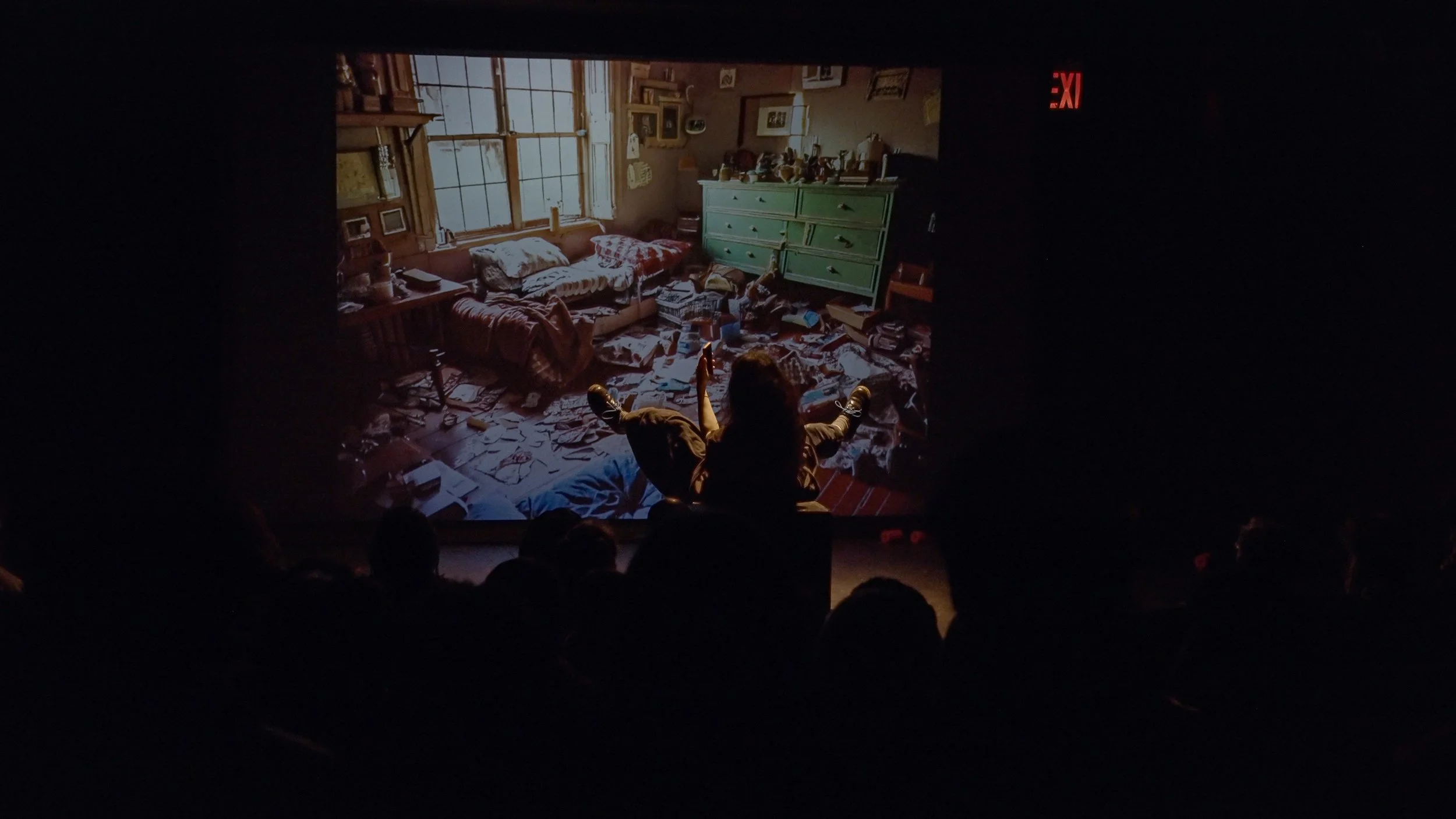 A person sitting with legs crossed and arms raised in front of a large screen displaying a cluttered, messy room with a window, a green dresser, and scattered papers and books.