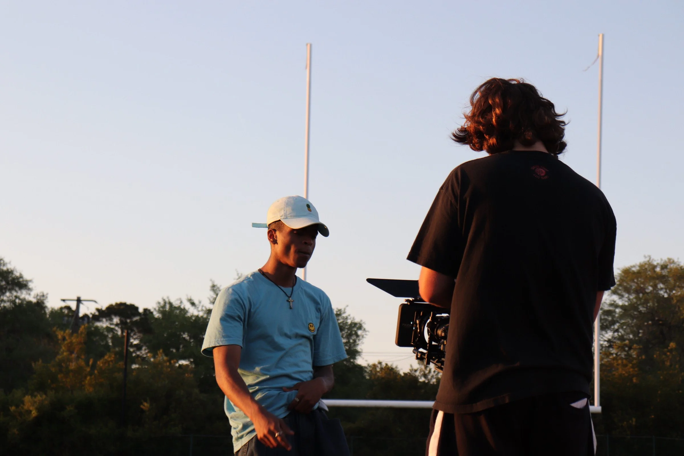 Two individuals standing on a football field, one wearing a white cap and light blue shirt, the other holding a camera, with football goalposts and trees in the background during sunset.