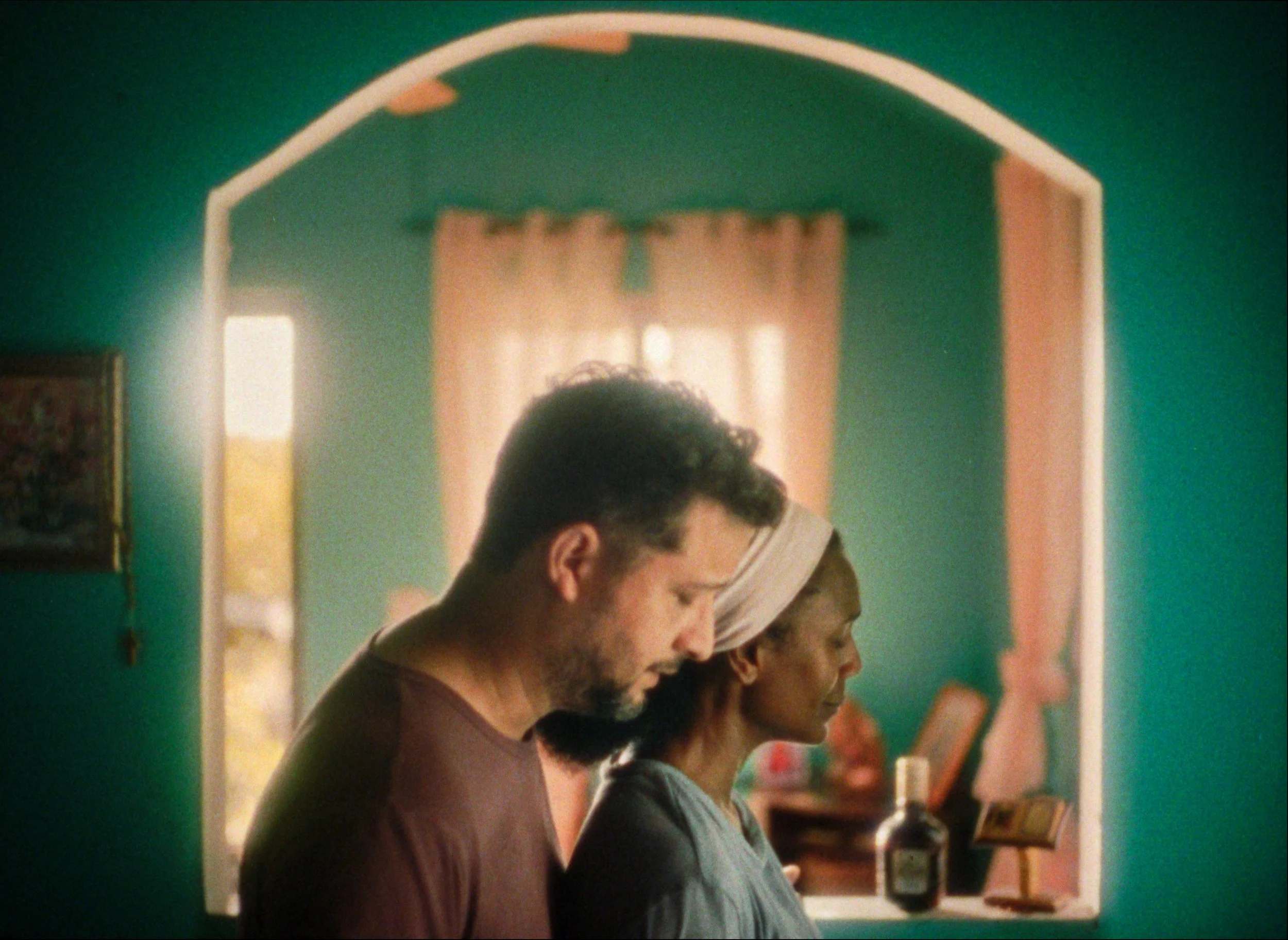 A man and woman stand side by side with their heads bowed in prayer or reflection inside a warmly lit room with teal walls, a window, and various personal items on the nearby shelves.