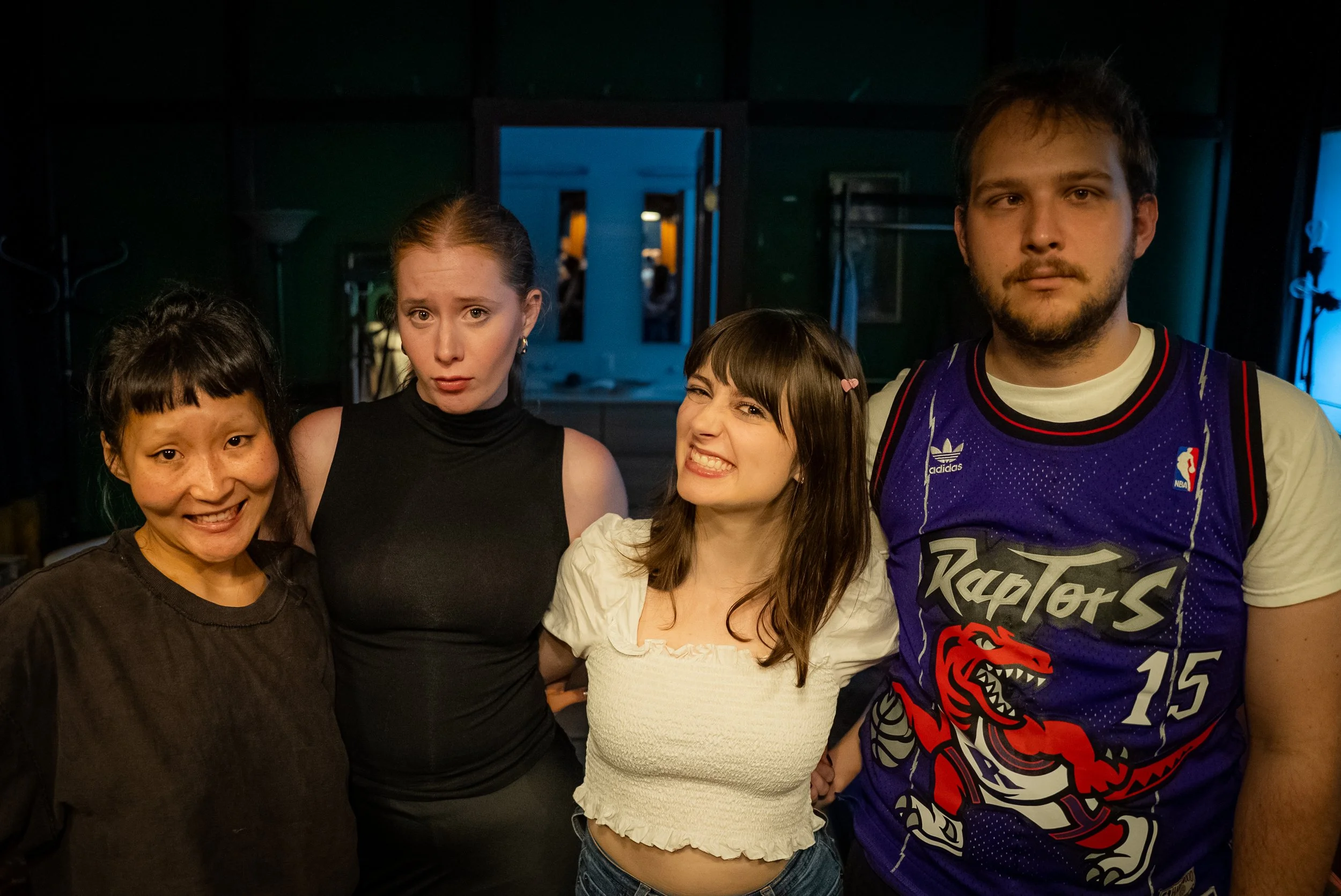 Four people standing close together in a dimly lit room, posing for a photo, with a mirror and cabinet in the background.
