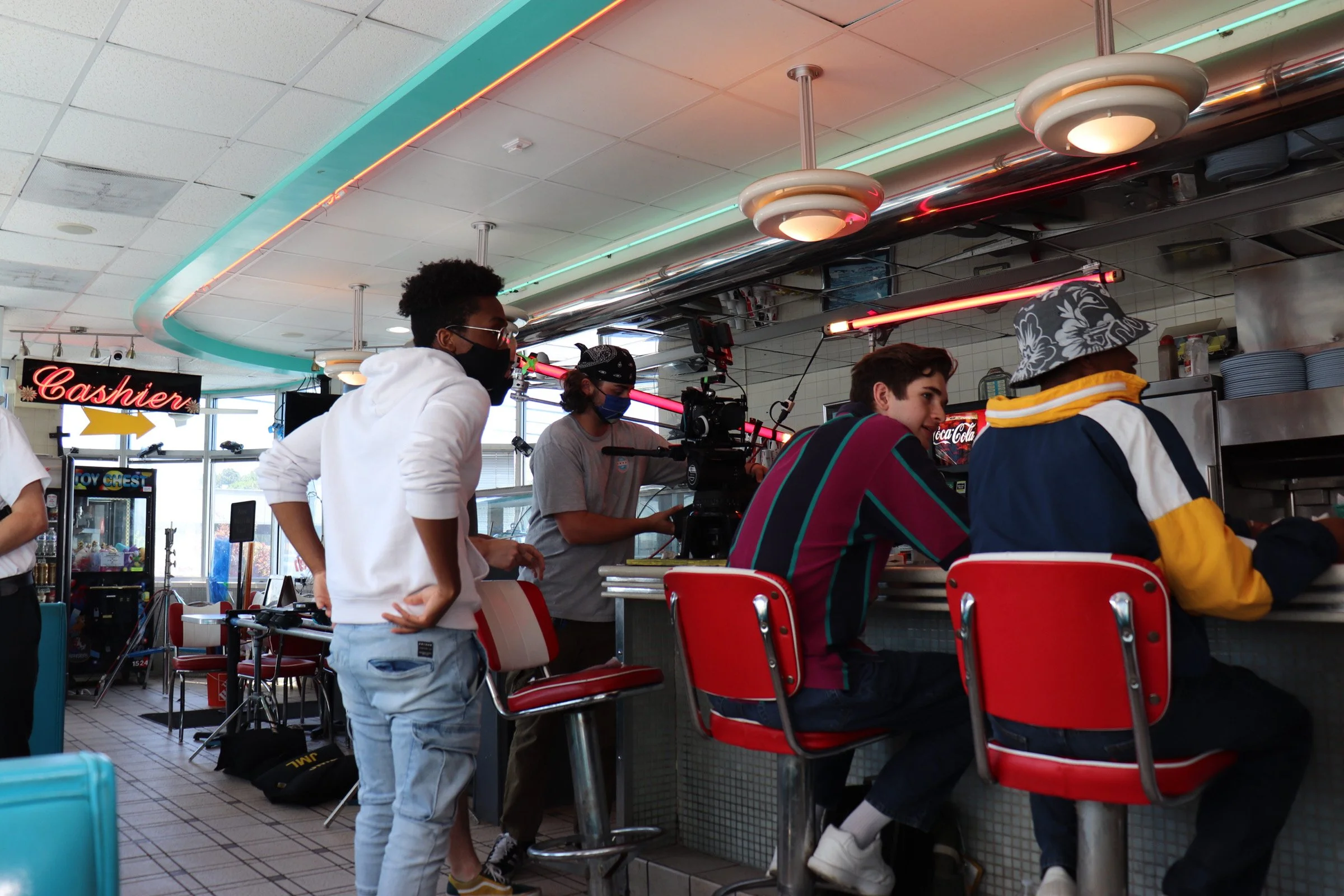 People filming a scene at a diner counter with a camera on a tripod, some sitting and talking, with a shier sign and neon lights overhead.