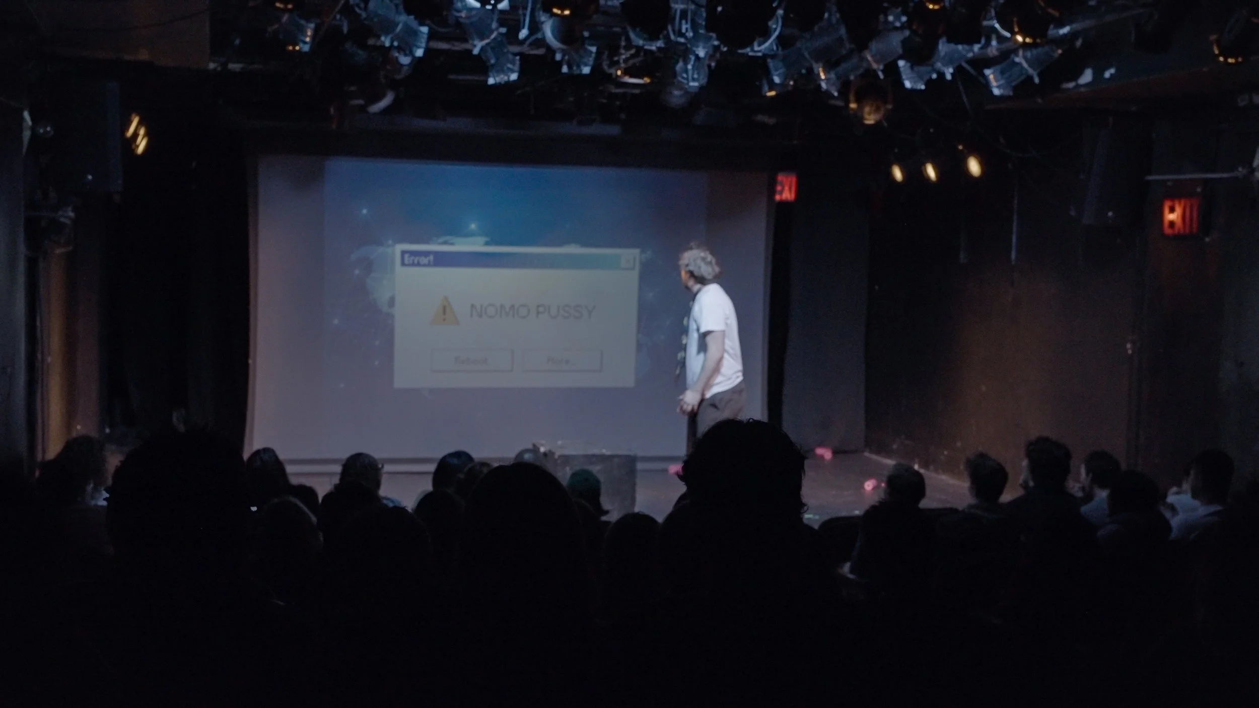A comedy show on stage with a man standing in front of a large screen displaying an error message that reads 'Error! NOMO PUSSY' and two buttons labeled 'Cancel' and 'Purse.'