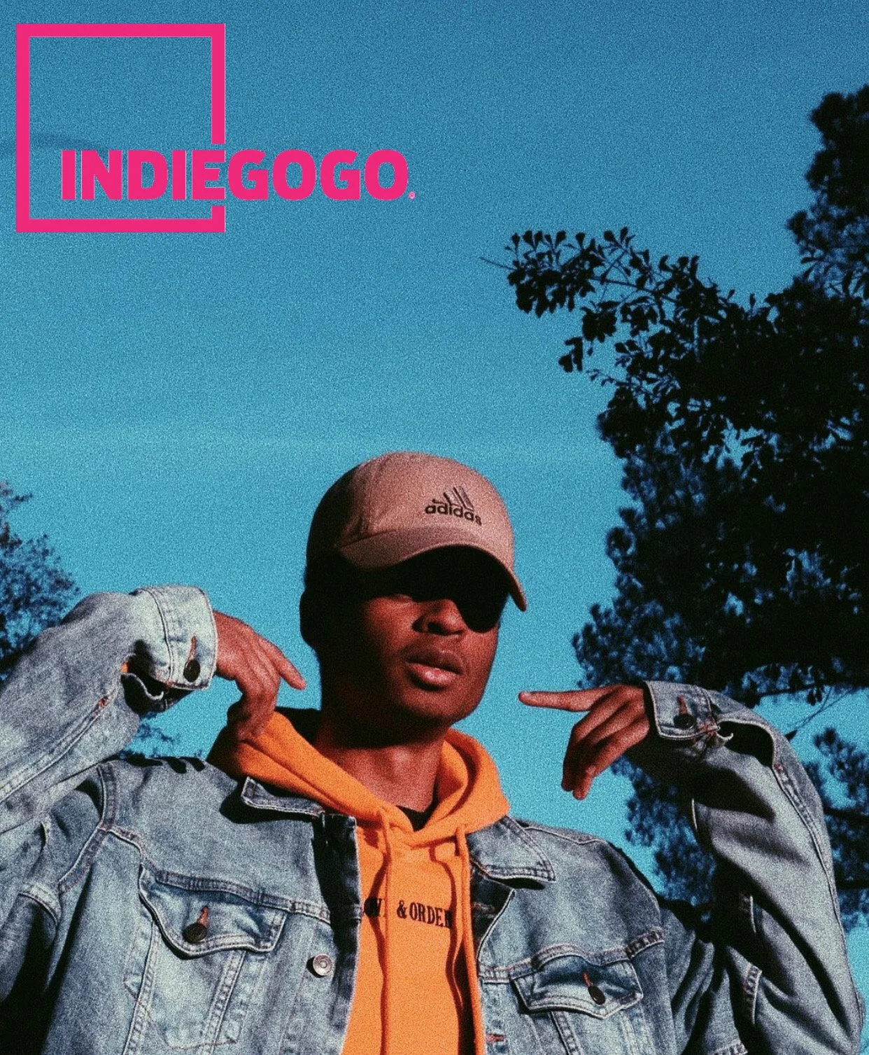 A person wearing a beige adidas cap, sunglasses, a denim jacket, and an orange hoodie, pointing at their head against a blue sky with trees in the background, with the 'IndieGogo' logo in the upper left corner.