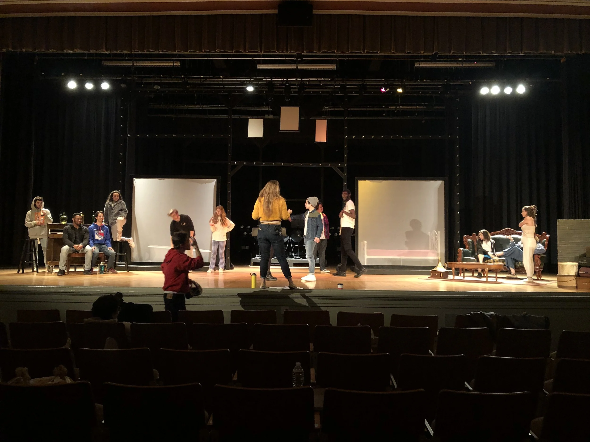 The stage of a theater with actors rehearsing and crew setting up, including people sitting, standing, and walking across the stage.
