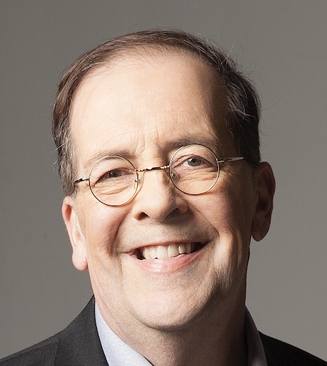 Close-up portrait of a smiling middle-aged man with glasses, wearing a dark suit and light-colored shirt.