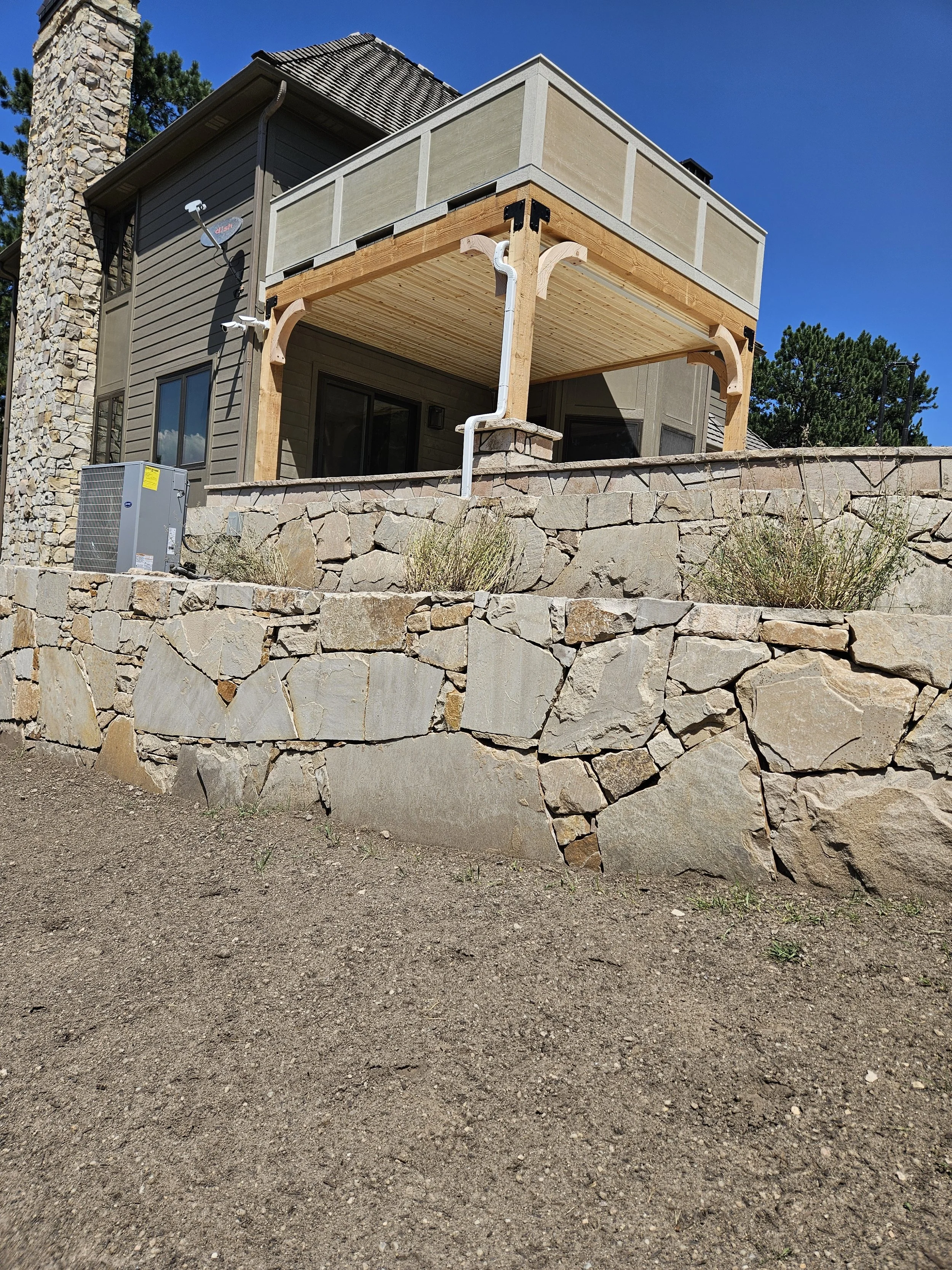 A house with a stone foundation, gray siding, and a chimney. There is a raised wooden deck with support columns, located behind a stone wall, with a dirt ground in the foreground.