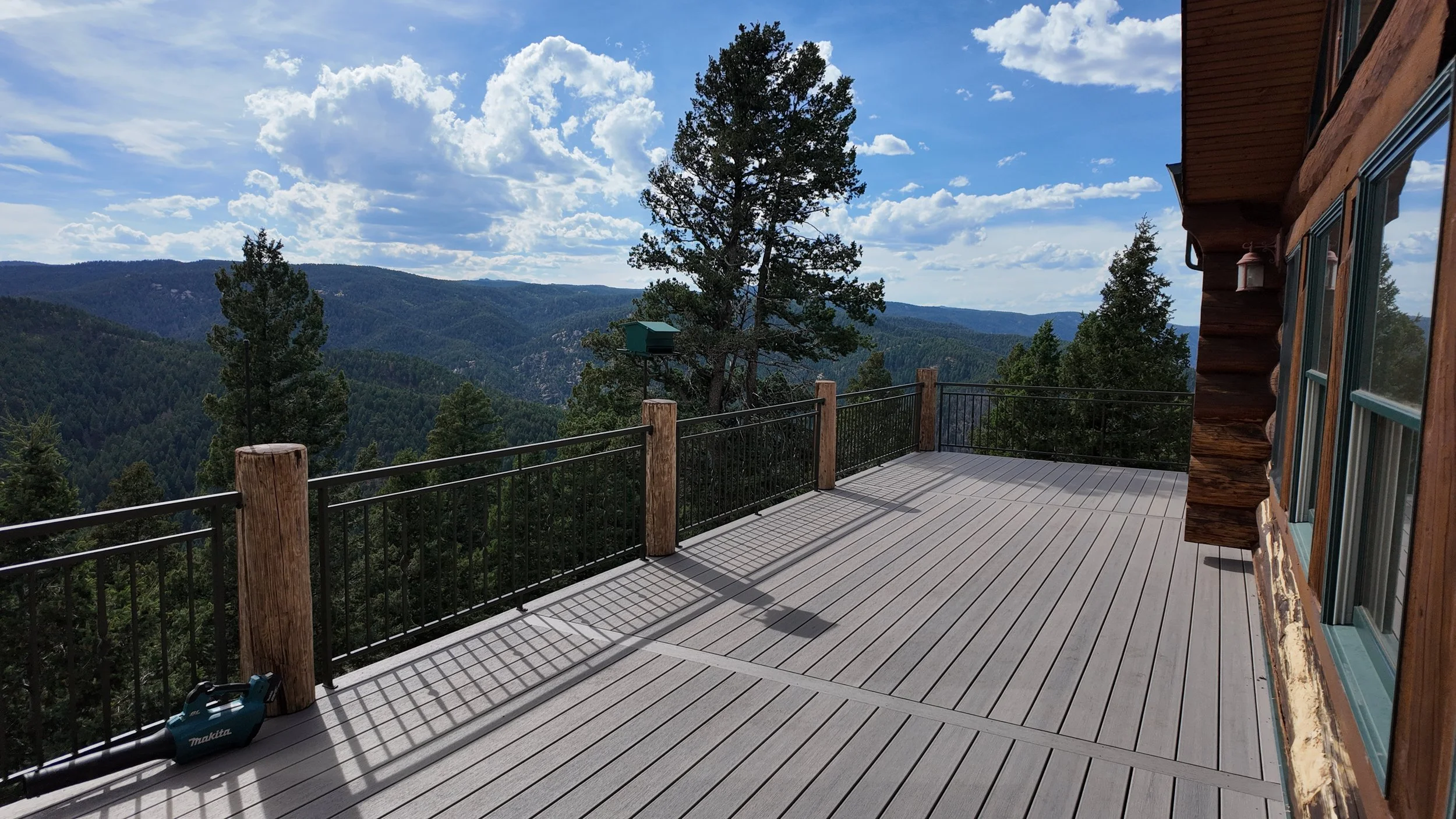 A spacious wooden deck on a mountain cabin with a black metal railing and large view of forested mountains under a partly cloudy sky.