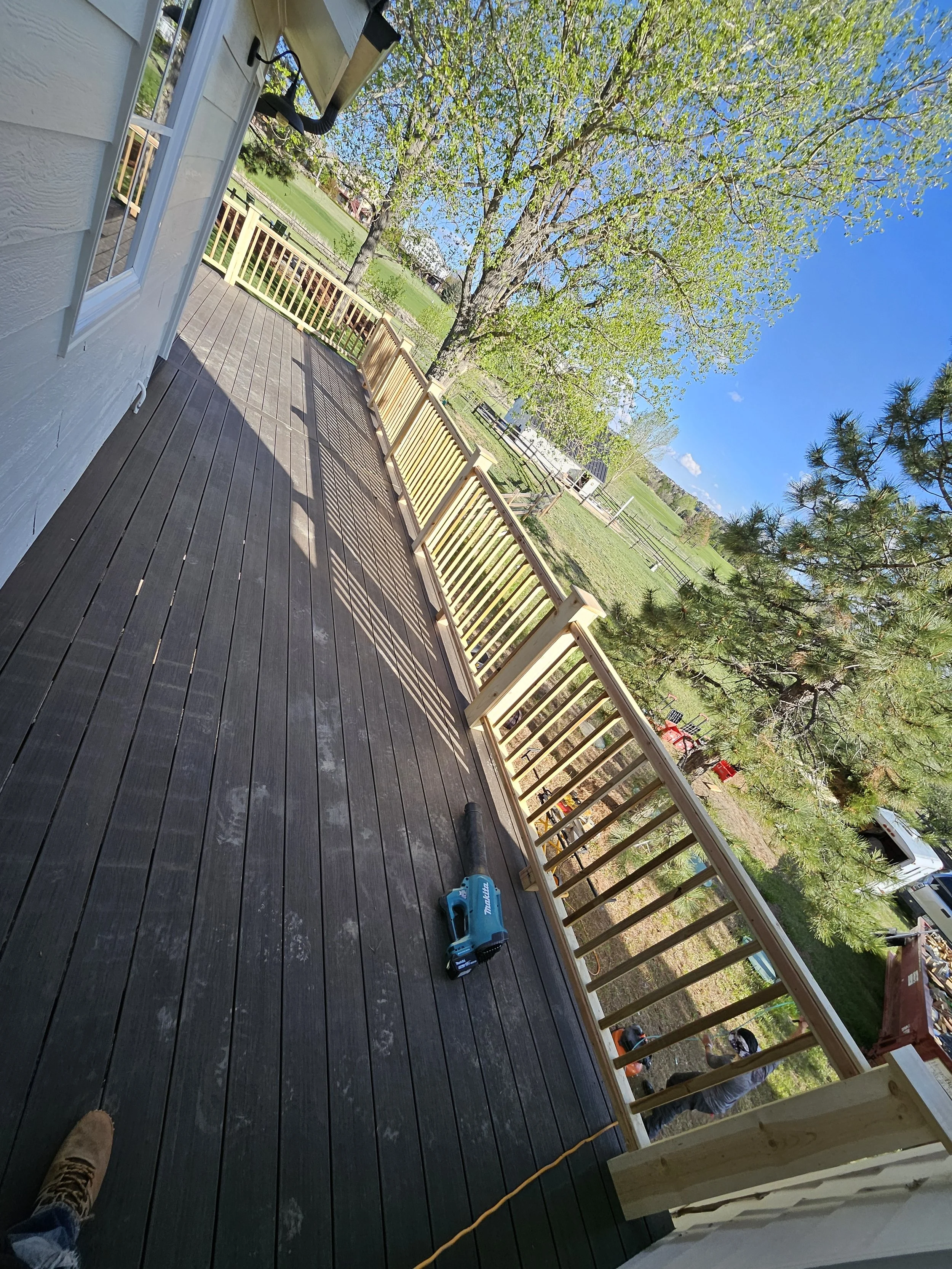 Construction of a wooden deck or porch with a new railing being built outside a white house on a sunny day. Tools are on the deck, and a person is working outside.