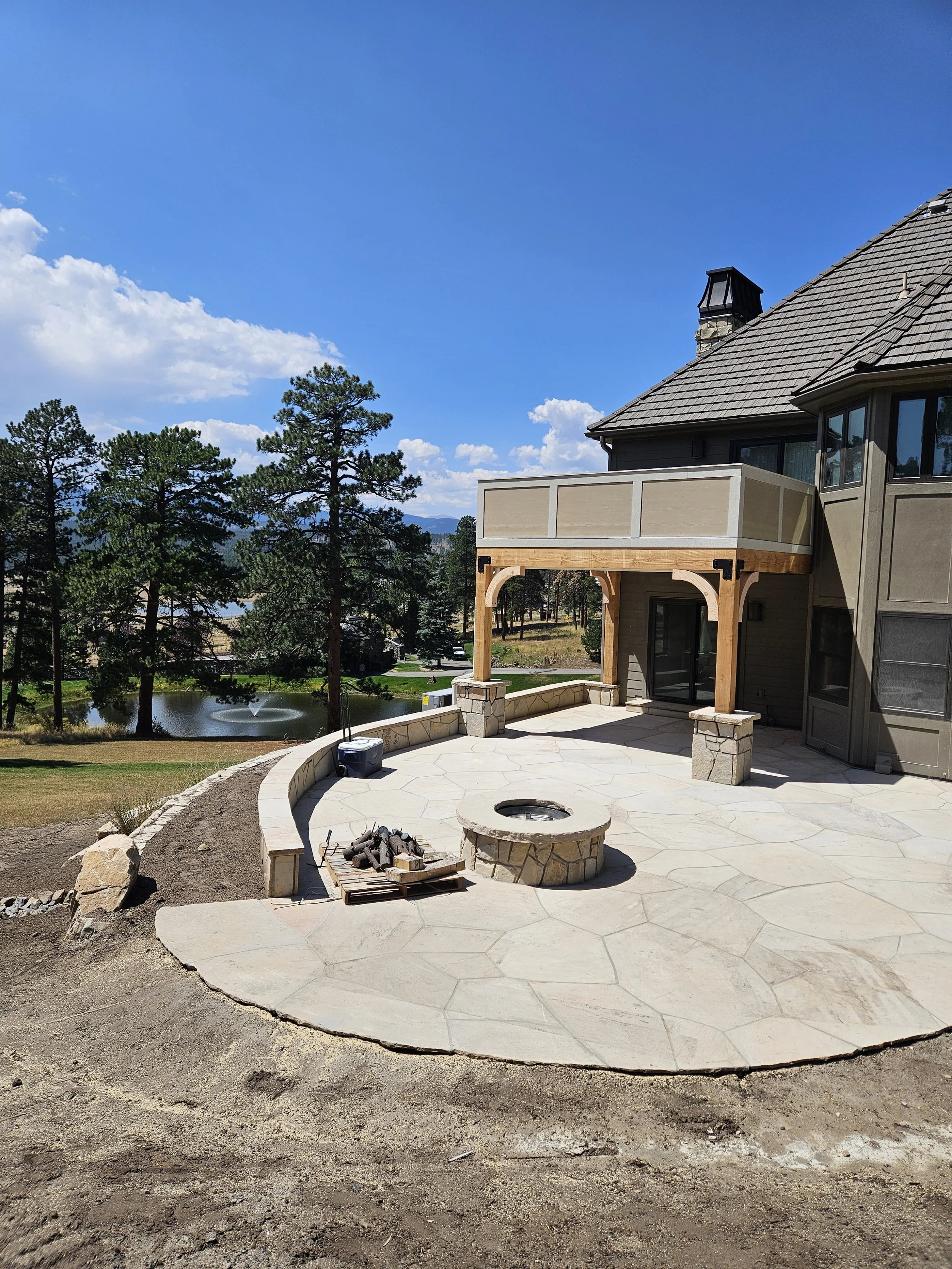Newly constructed stone patio with fire pit, part of a backyard, with trees and a pond in the background, under a bright blue sky.