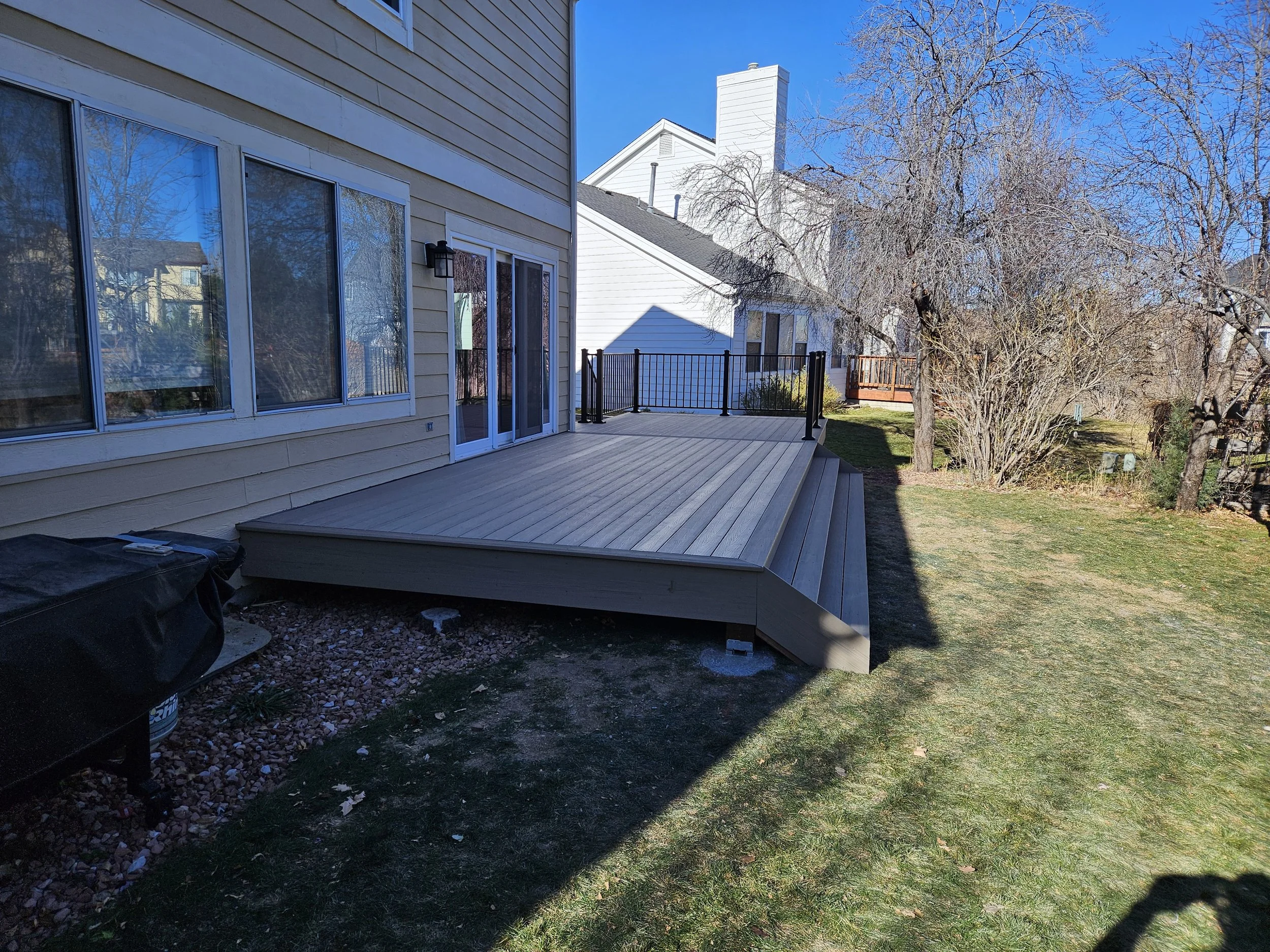 Image of a new wooden deck attached to beige house with sliding glass doors, black railing, and a grassy backyard with trees and neighboring houses in the background.
