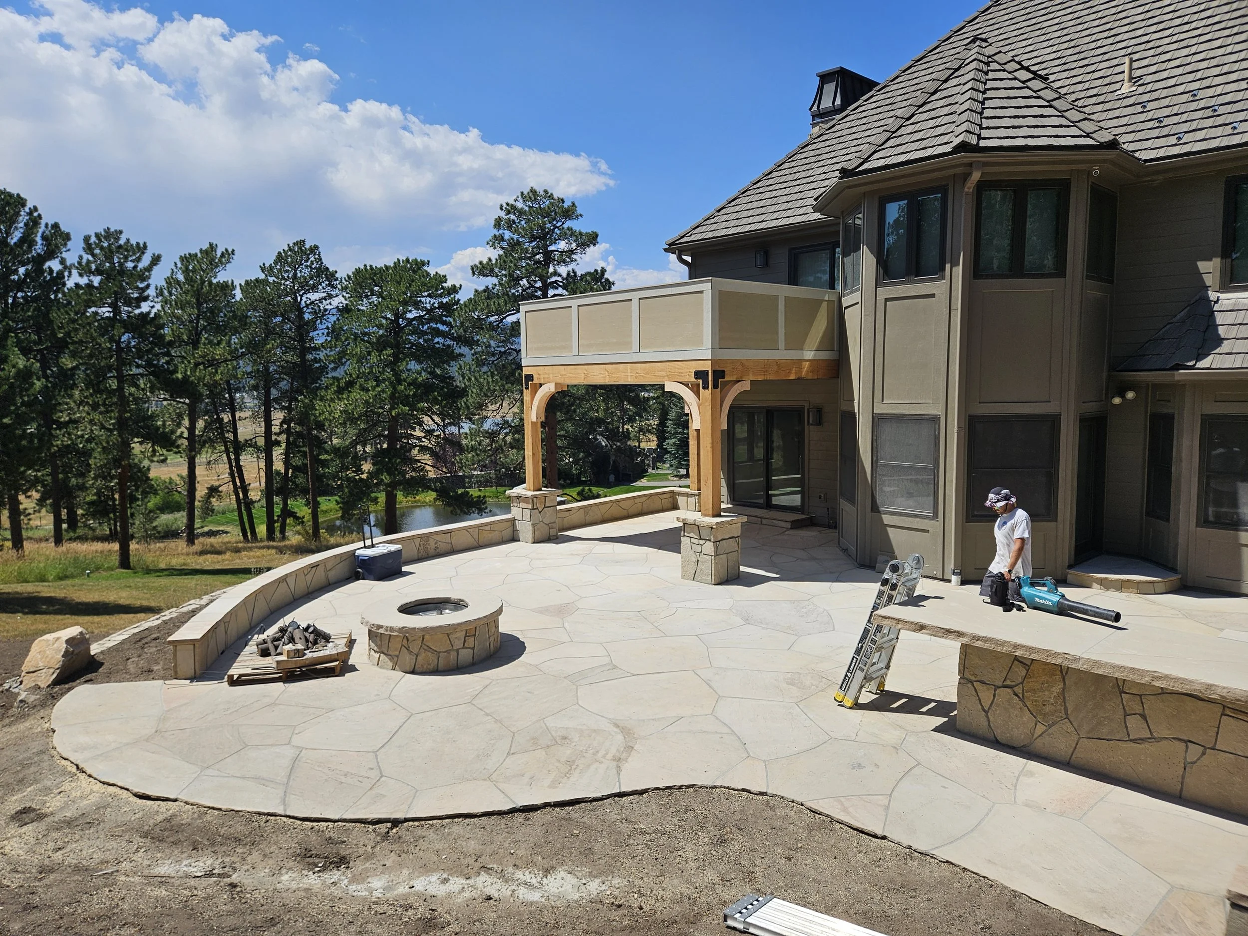 Residential backyard patio under construction with stone fire pit, wooden pergola, and building work in progress. A person wearing white shirt and hat stands on the patio.