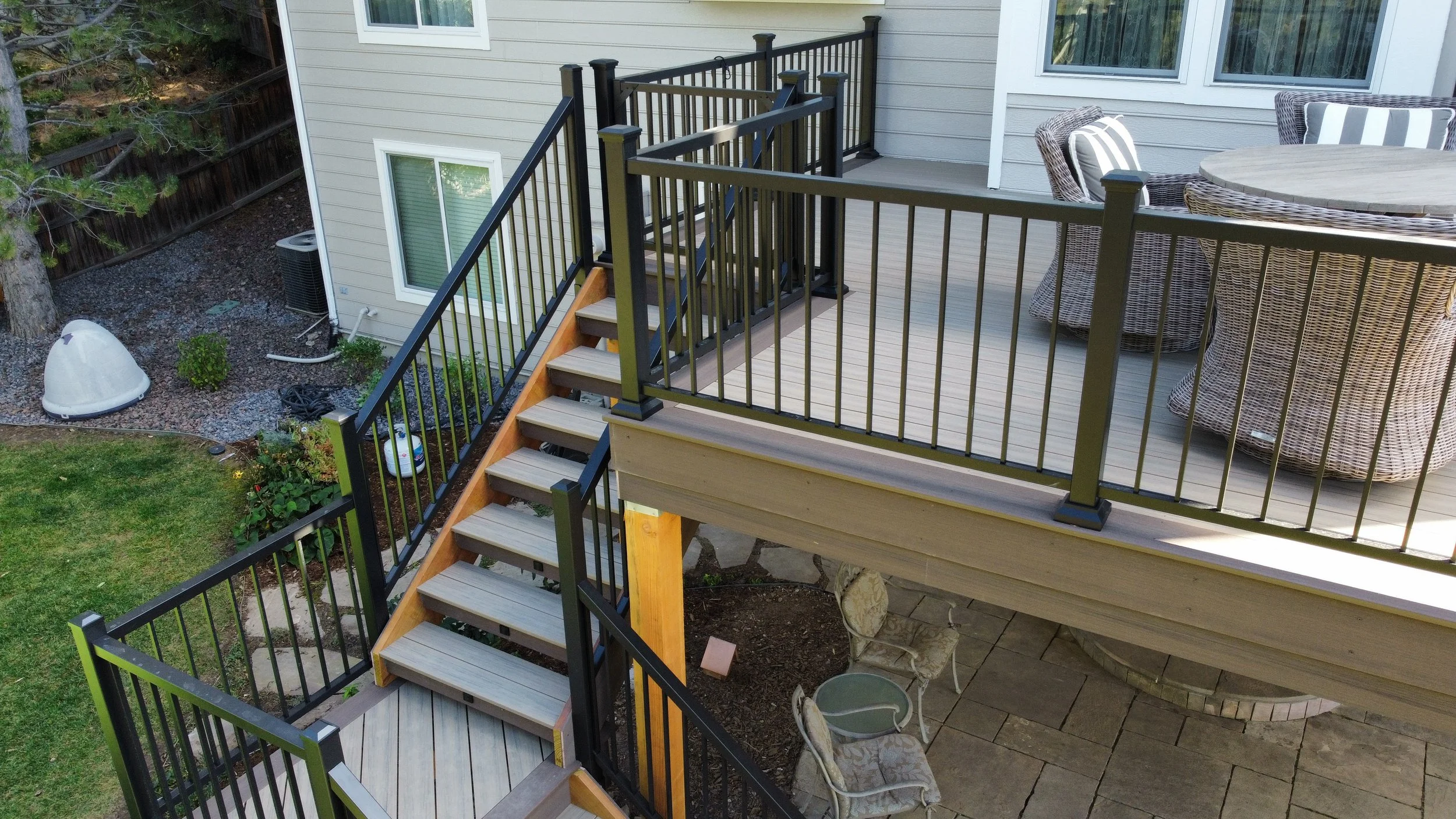 A multi-level outdoor wooden deck with black metal railings, outdoor chairs, a round table, and stairs leading down to a yard with green grass and a paved patio area.
