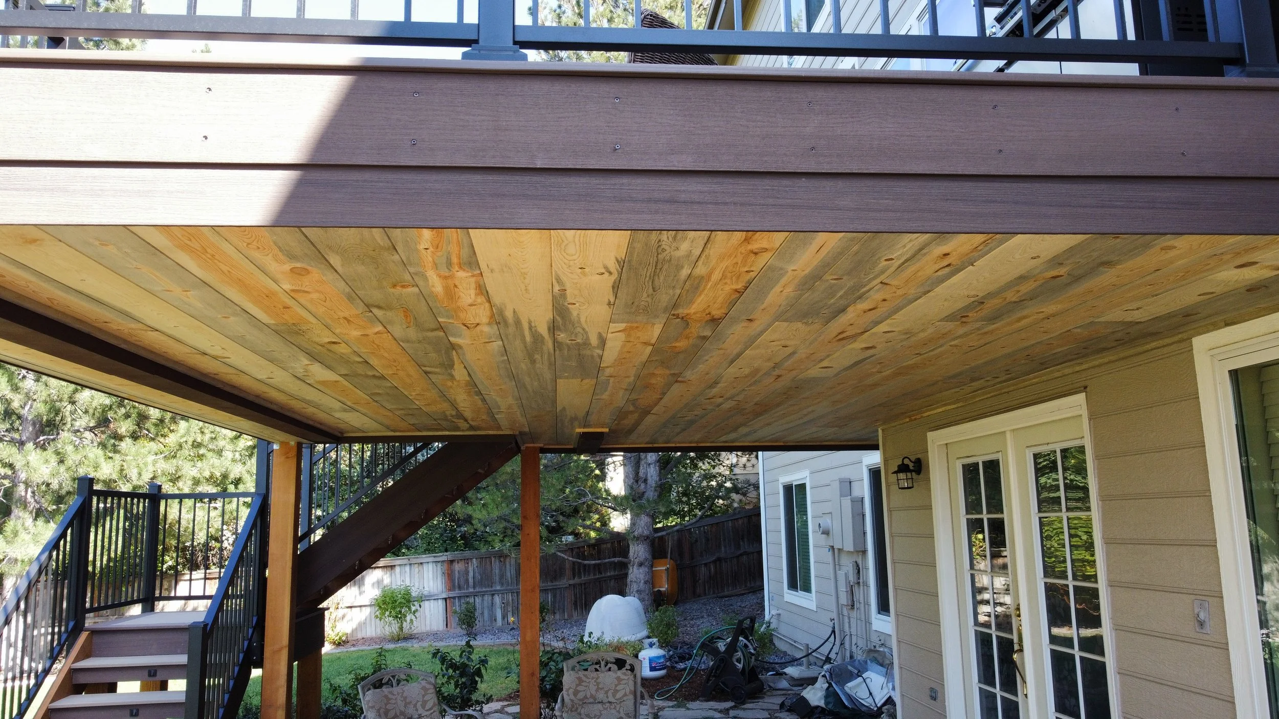 View of a house's backyard with a newly constructed wooden deck overhead, exposed wood ceiling, stairs, and fencing, with outdoor furniture and gardening tools.