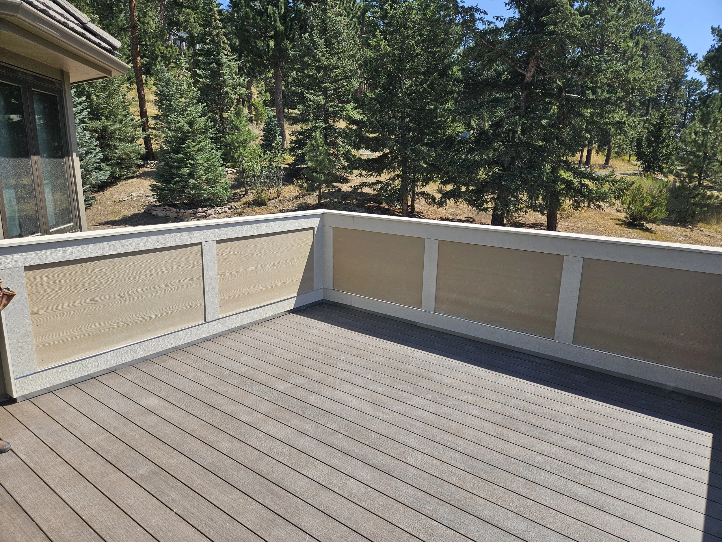 Empty outdoor wooden deck with a beige and white railing, overlooking a forested area of tall green pine trees and a clear blue sky.